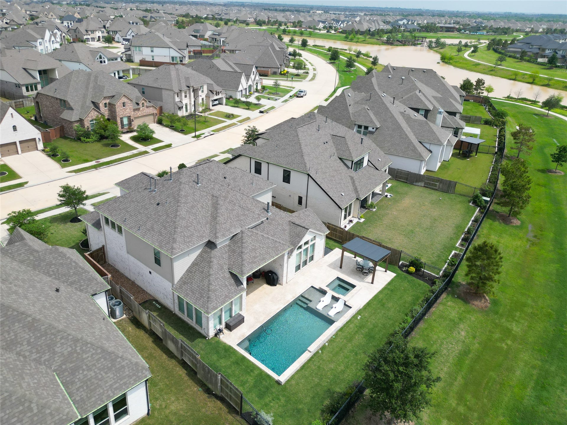 A modern suburban neighborhood with spacious homes. The featured property includes a backyard with a swimming pool, spa, and a covered patio area, set in a well-maintained yard. The neighborhood is characterized by neatly lined houses, green spaces, and nearby water features, offering a serene and inviting environment.