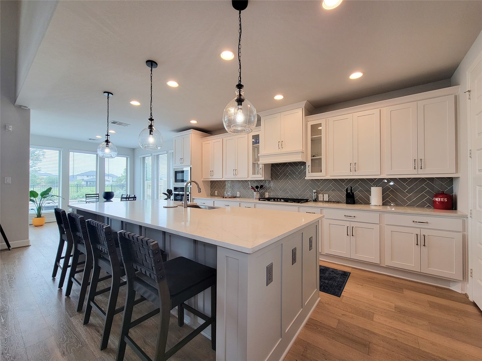 This modern kitchen boasts a spacious layout with a large island featuring seating for four. It includes sleek white cabinetry, stainless steel appliances, and stylish pendant lighting, all enhanced by ample natural light from the adjoining dining area.