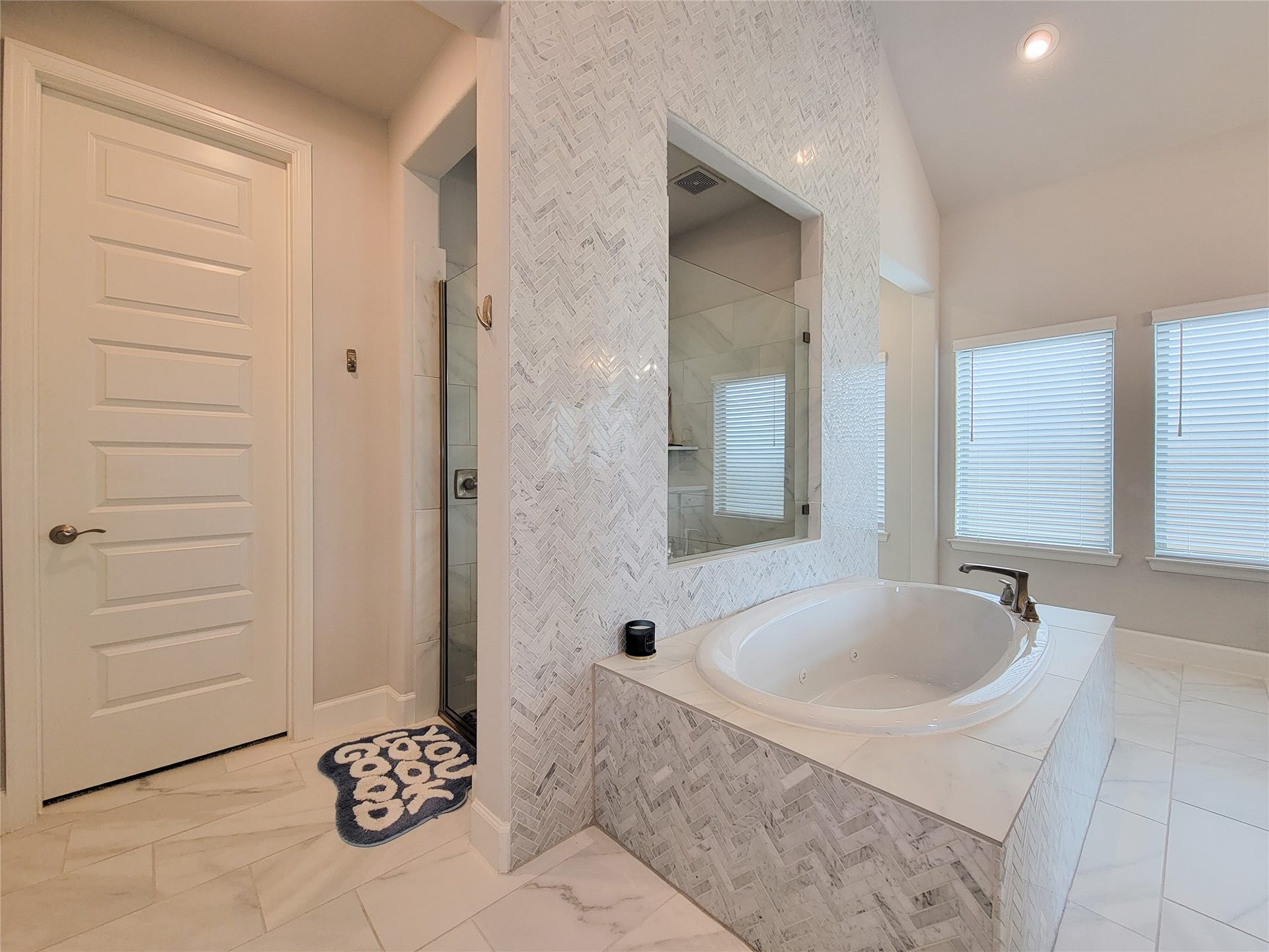 This bathroom features a luxurious, large soaking tub set in a marble-tiled surround, a separate glass-enclosed shower, and ample natural light from two windows with blinds, creating a bright and relaxing space.