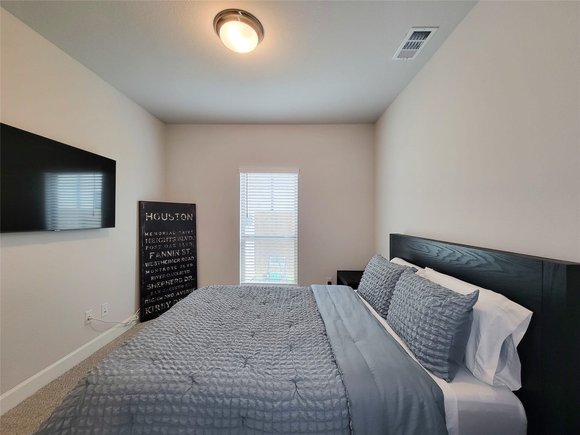 This cozy bedroom features a large window allowing natural light. The room has a simple, modern design with neutral walls and a ceiling light fixture.