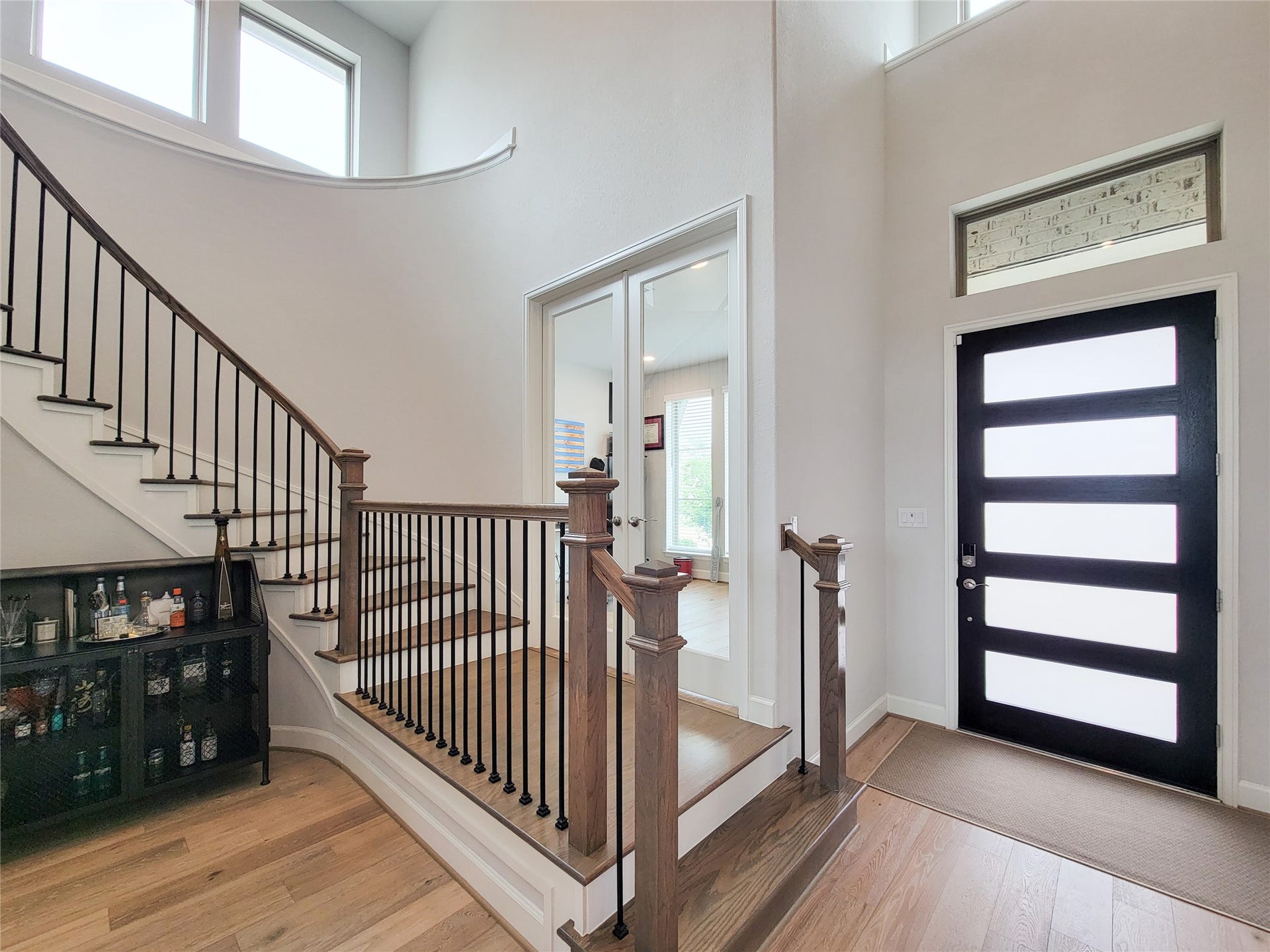 This photo showcases a modern entryway with a sleek black front door featuring frosted glass panels. The space is bright, with natural light streaming in from high windows. A staircase with wooden steps and iron railings leads to the upper floor, while a small bar area sits below. The design is open and inviting, with light wood flooring adding warmth.