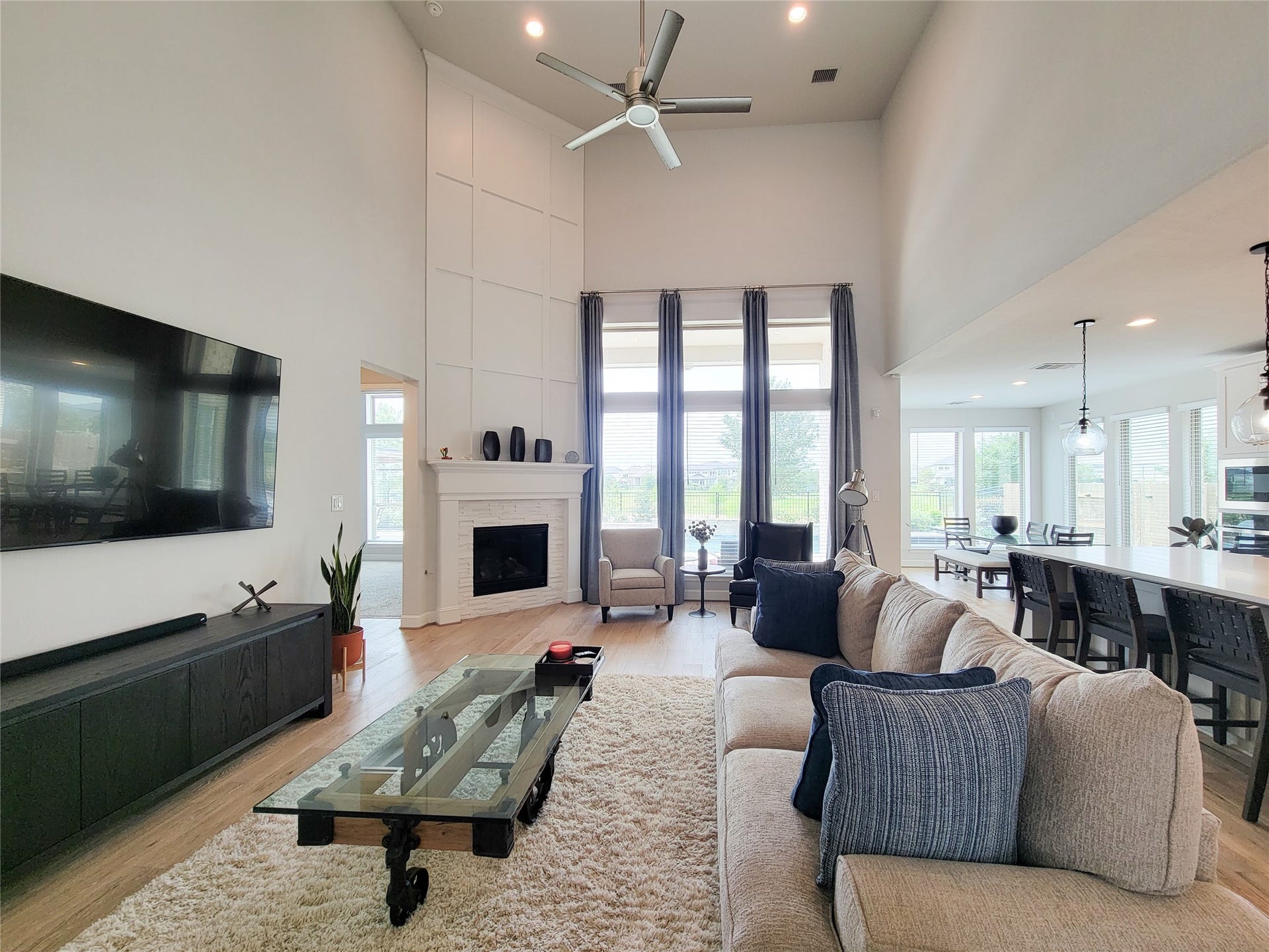 This spacious living room features high ceilings, a modern ceiling fan, and a large fireplace. Natural light pours in through tall windows, highlighting the open layout that seamlessly connects to the dining and kitchen areas.