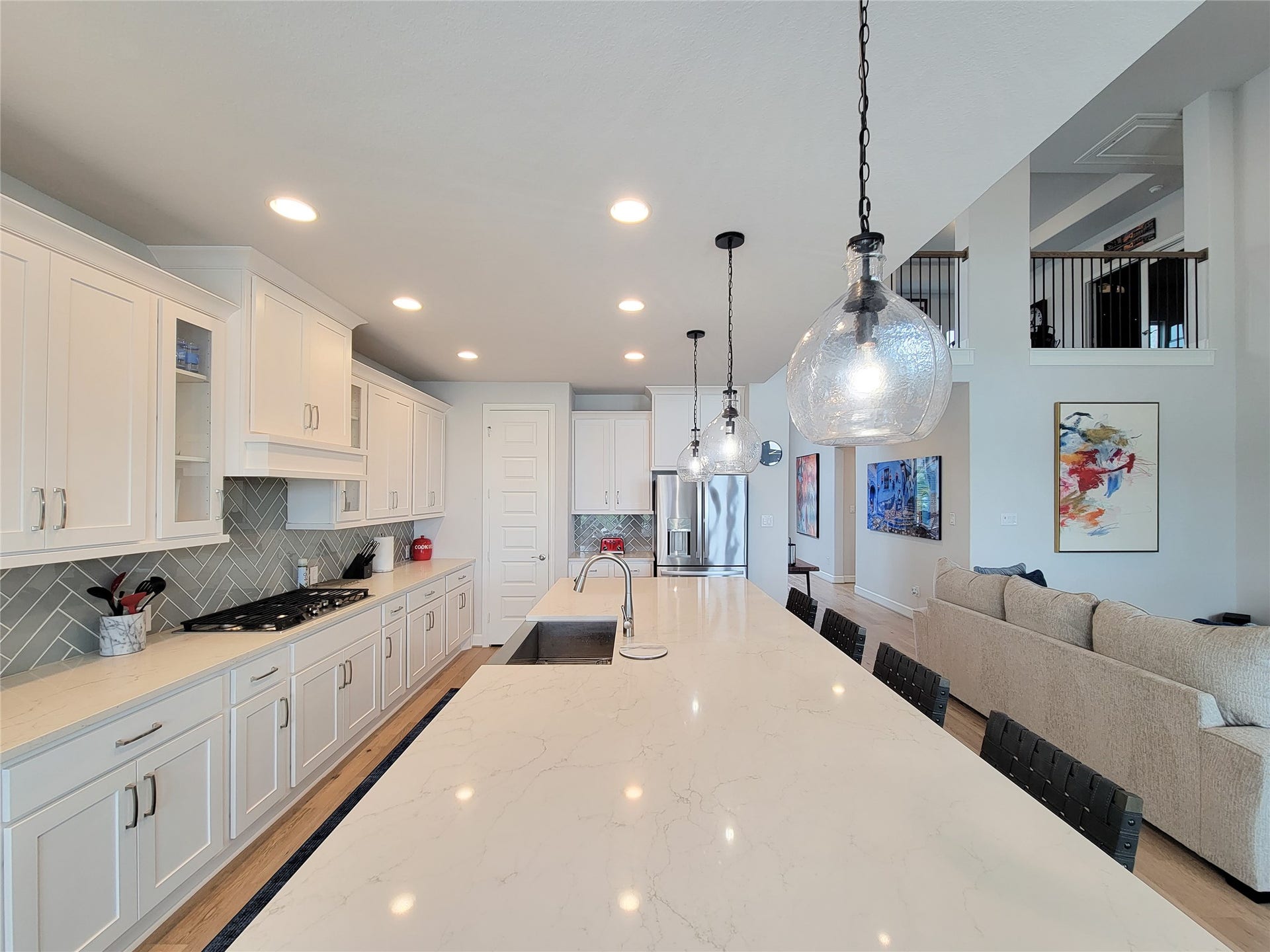 This spacious kitchen features a long marble island with pendant lighting, white cabinetry, stainless steel appliances, and an open layout leading into a living area with modern decor and high ceilings.