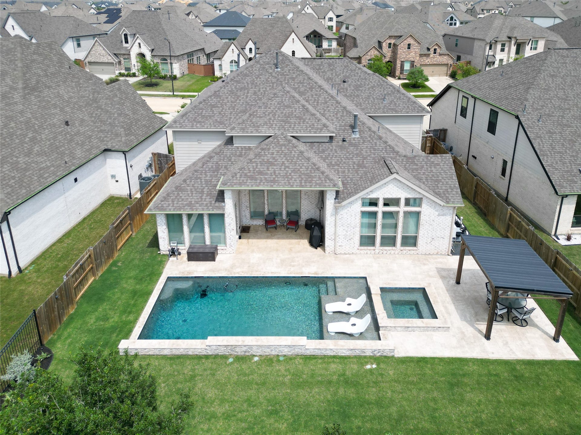 This aerial view showcases a modern home with a spacious backyard featuring a pool and spa. The property includes a covered patio area and is surrounded by a well-maintained lawn and privacy fences, set in a neighborhood with similar style homes.