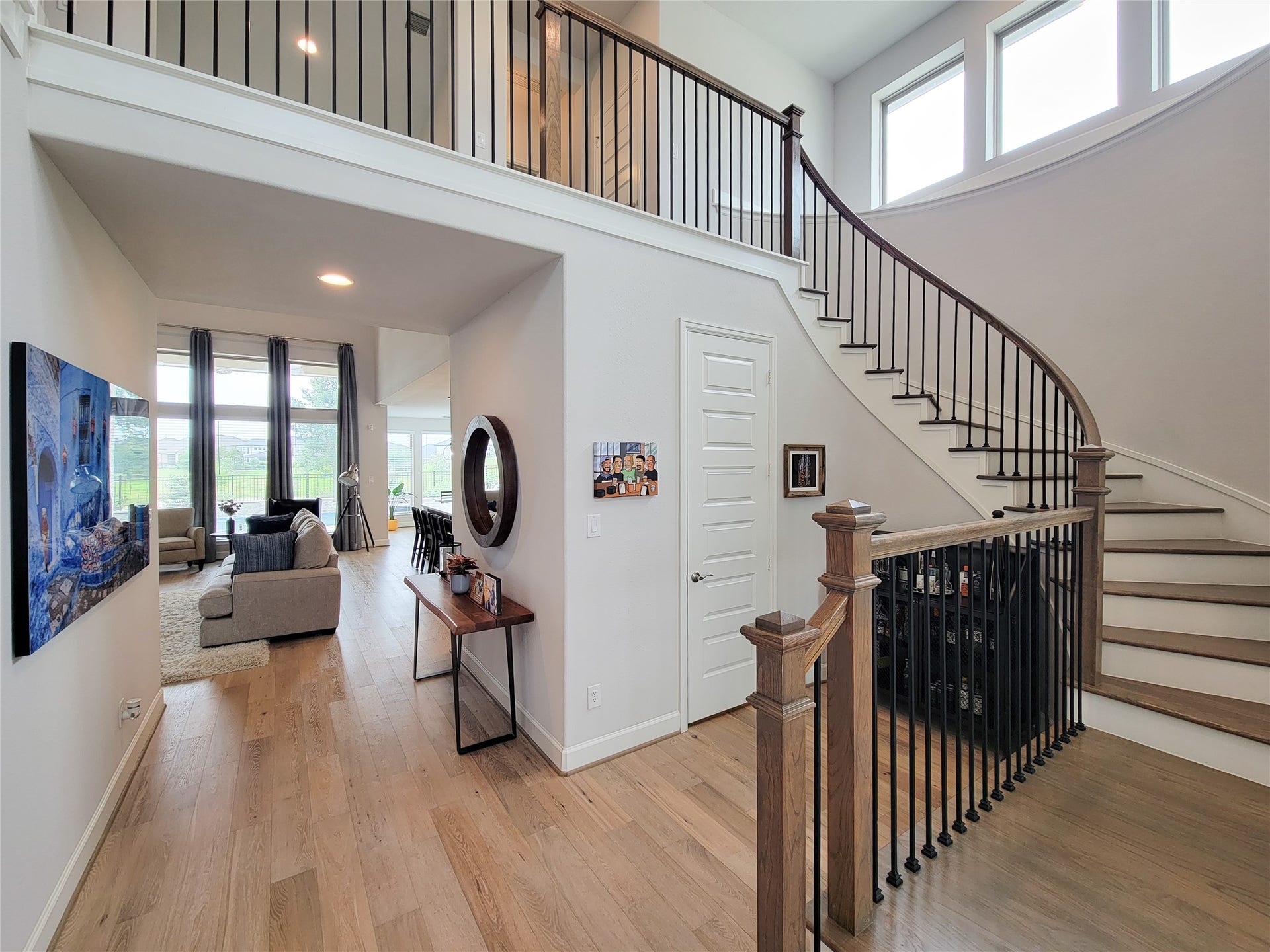 Bright, open entryway with a stunning curved staircase and modern railing. The space features light wood flooring and high ceilings, leading to a spacious living area with large windows and natural light.