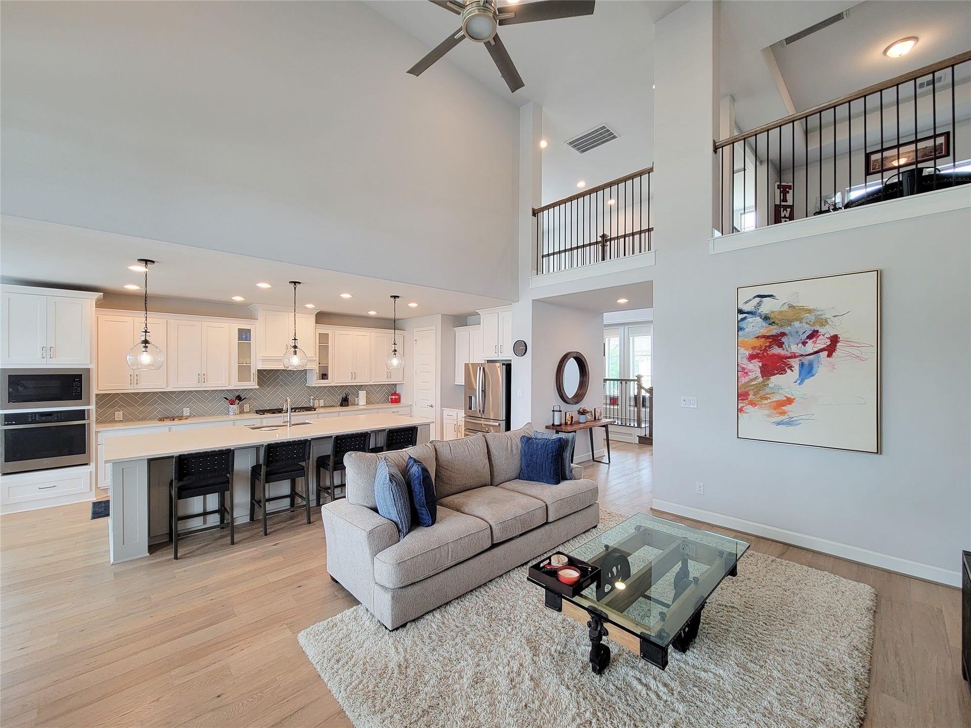 This open-concept living space features high ceilings, a modern kitchen with white cabinetry and pendant lighting, and a cozy seating area. A spacious loft overlooks the area, adding to the airy feel.