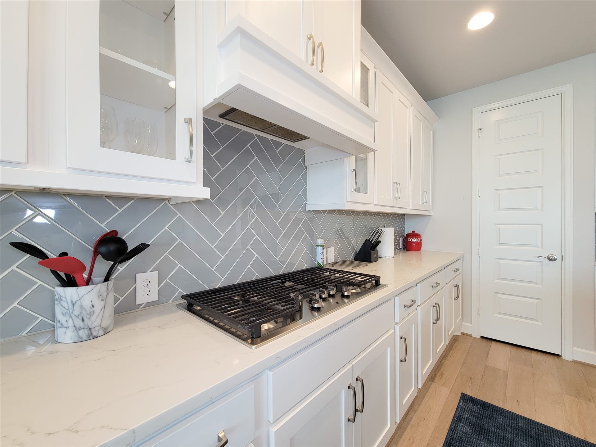 This kitchen features sleek white cabinetry with modern hardware, a stylish herringbone backsplash, and a spacious countertop. The built-in gas cooktop offers functionality, while the natural light enhances the bright, airy feel.