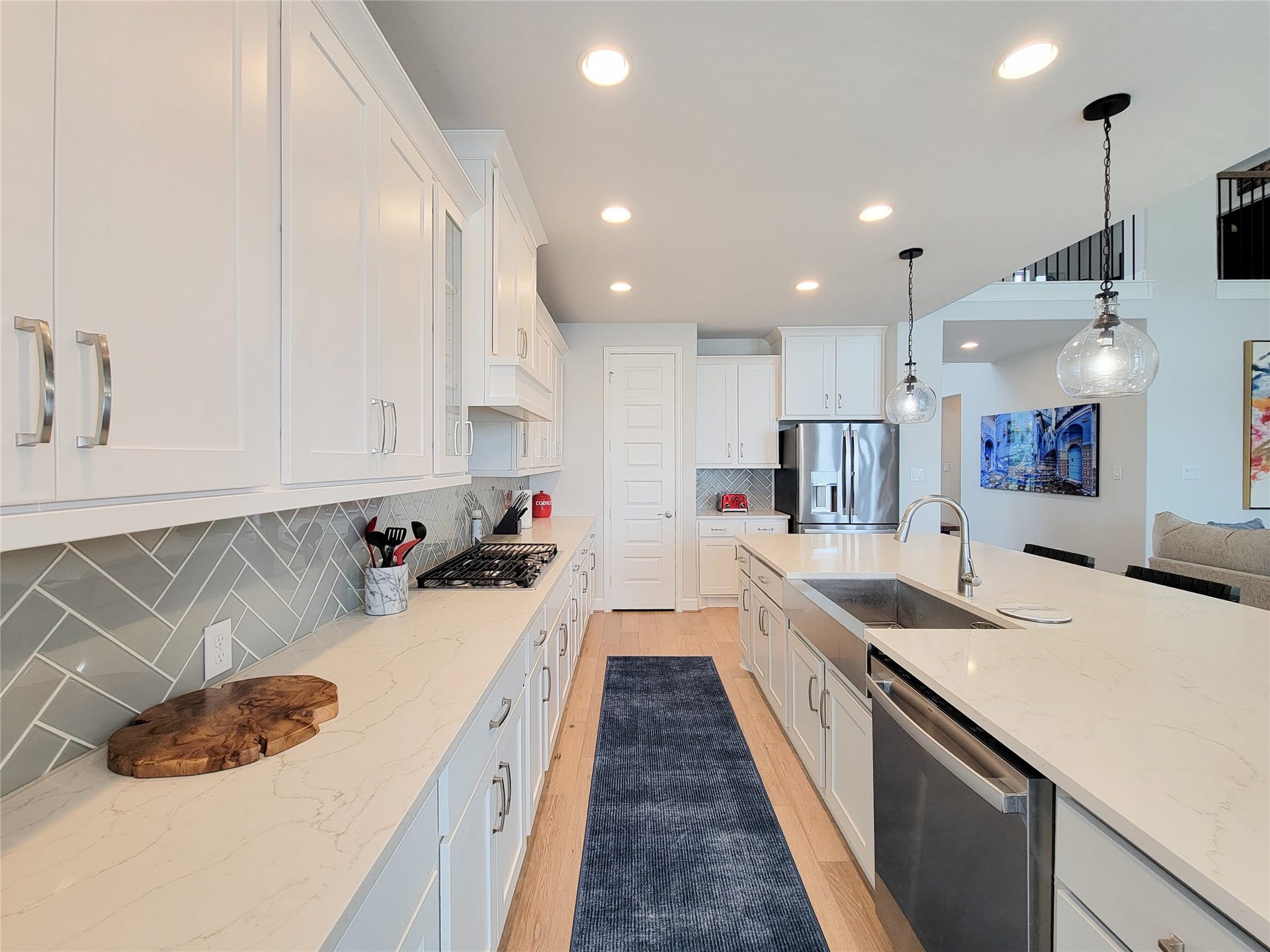 This modern kitchen features sleek white cabinetry, a spacious island with a sink, and stainless steel appliances. The herringbone backsplash adds a stylish touch, while pendant lighting creates a warm ambiance. Perfect for cooking and entertaining.