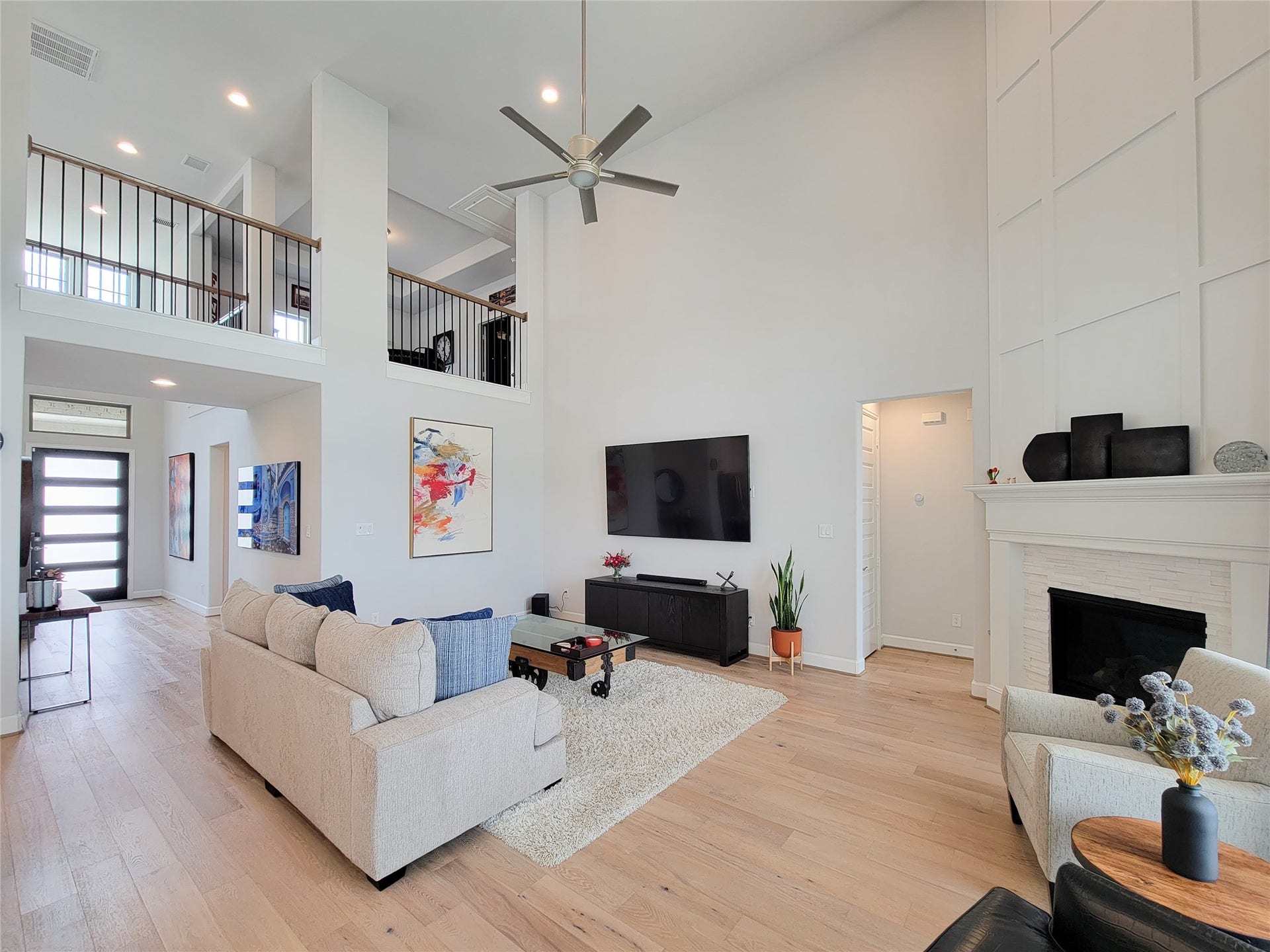 This spacious living room features high ceilings, a modern ceiling fan, and abundant natural light. It includes a cozy fireplace and an open loft area. The neutral color palette, hardwood floors, and contemporary decor create a welcoming atmosphere.