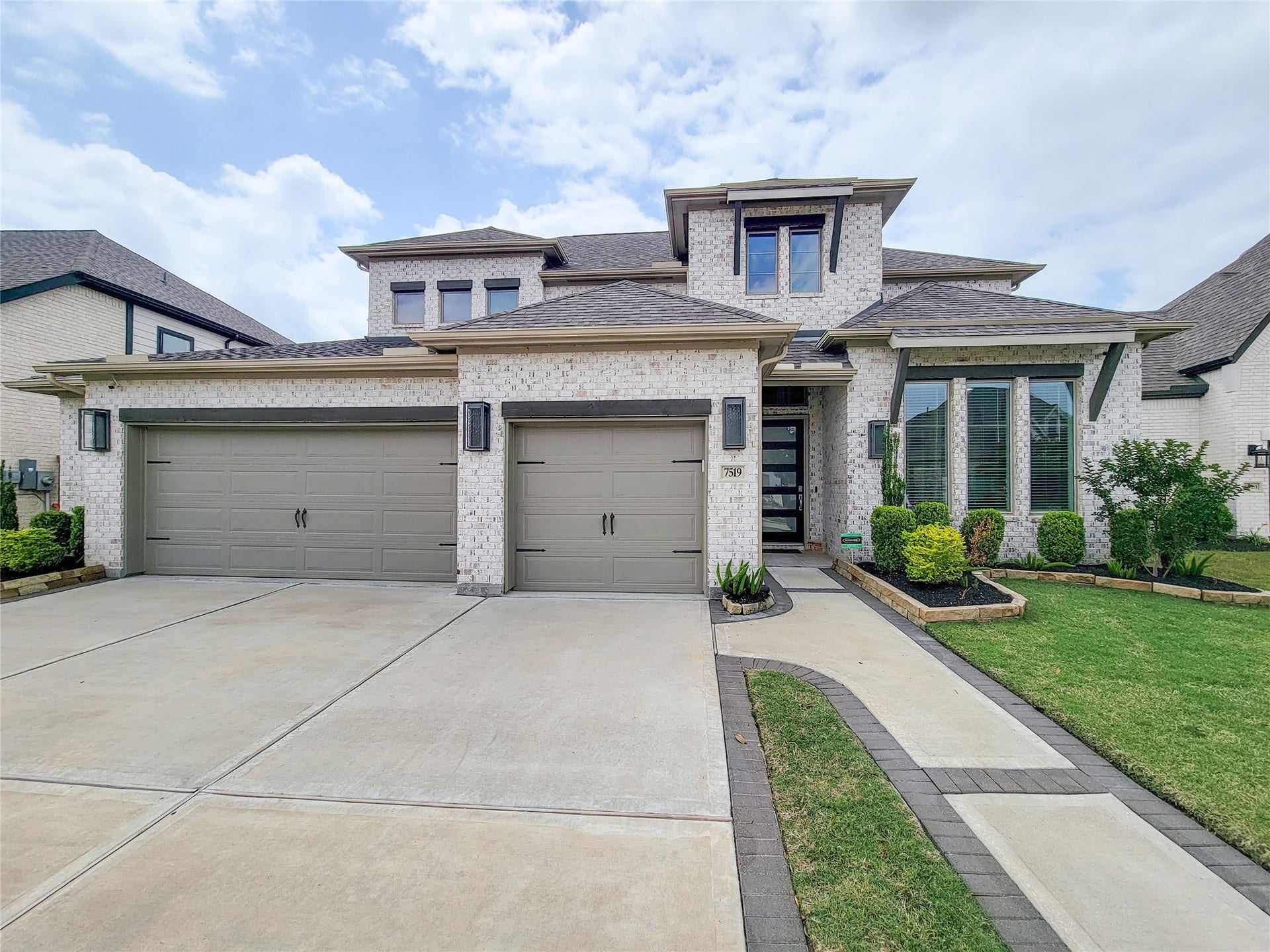This modern two-story home features a large three-car garage, elegant brick exterior, and well-maintained landscaping. The front entrance is inviting with contemporary design elements and ample windows for natural light.