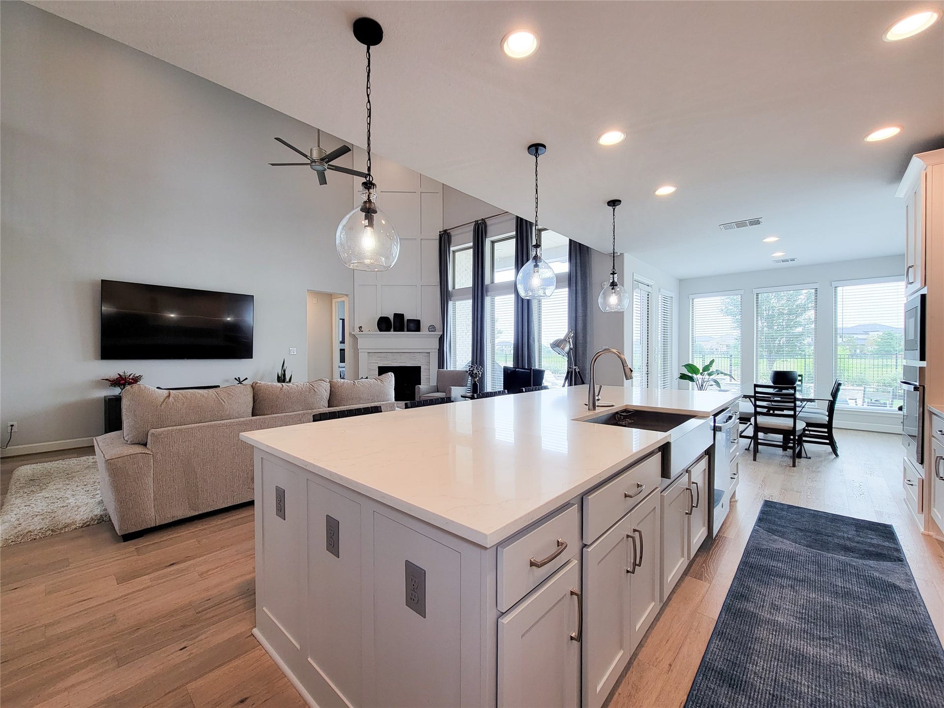 This open-concept living space features a modern kitchen with a large island, pendant lighting, and ample cabinetry. The area flows seamlessly into a comfortable living room with a mounted TV, cozy fireplace, and large windows offering natural light and views. Perfect for entertaining and family gatherings.