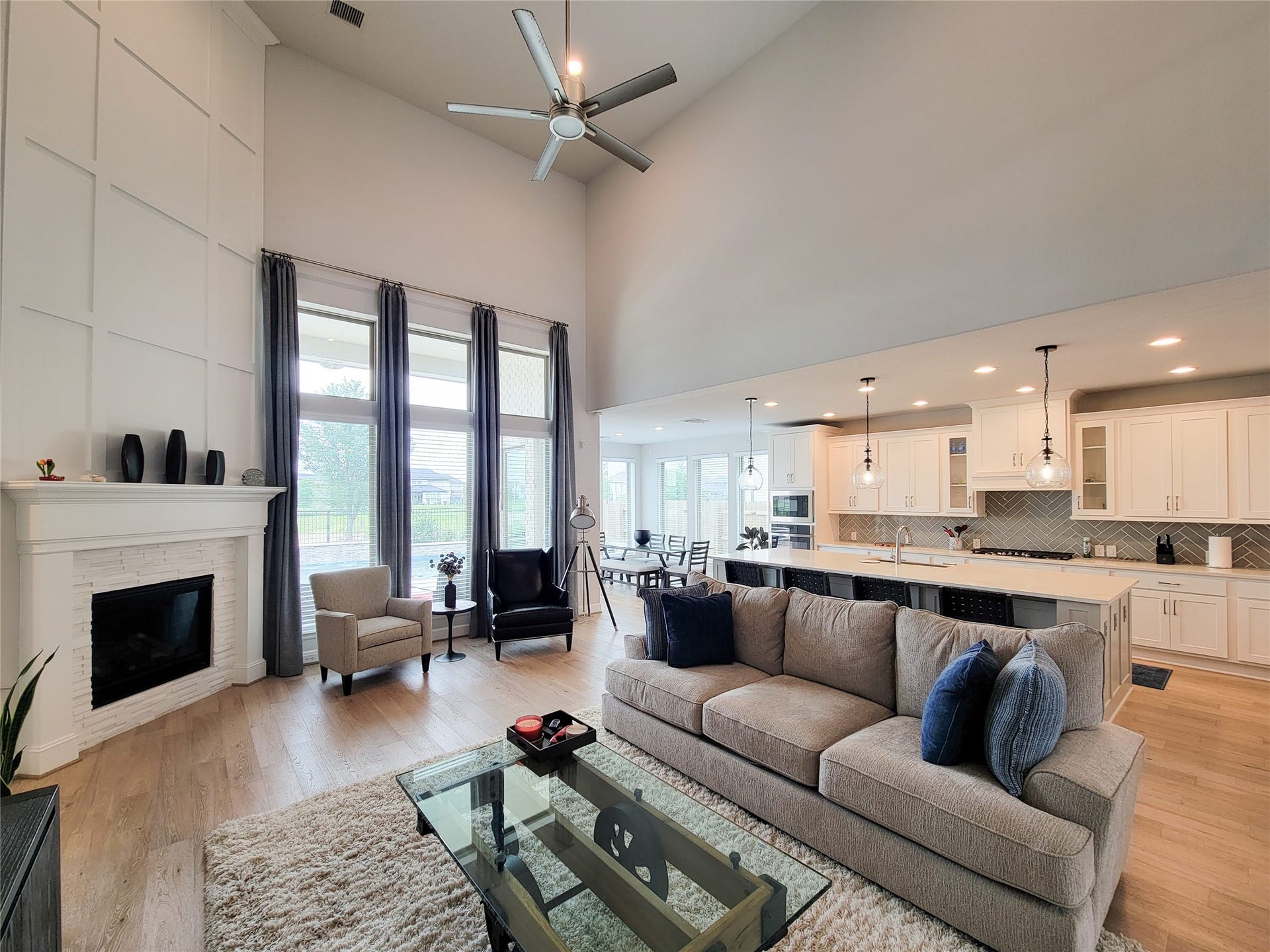 Spacious open-concept living area with high ceilings and large windows, featuring a cozy fireplace, modern kitchen with ample cabinetry, and a large island with seating. Natural light enhances the warm, inviting atmosphere.