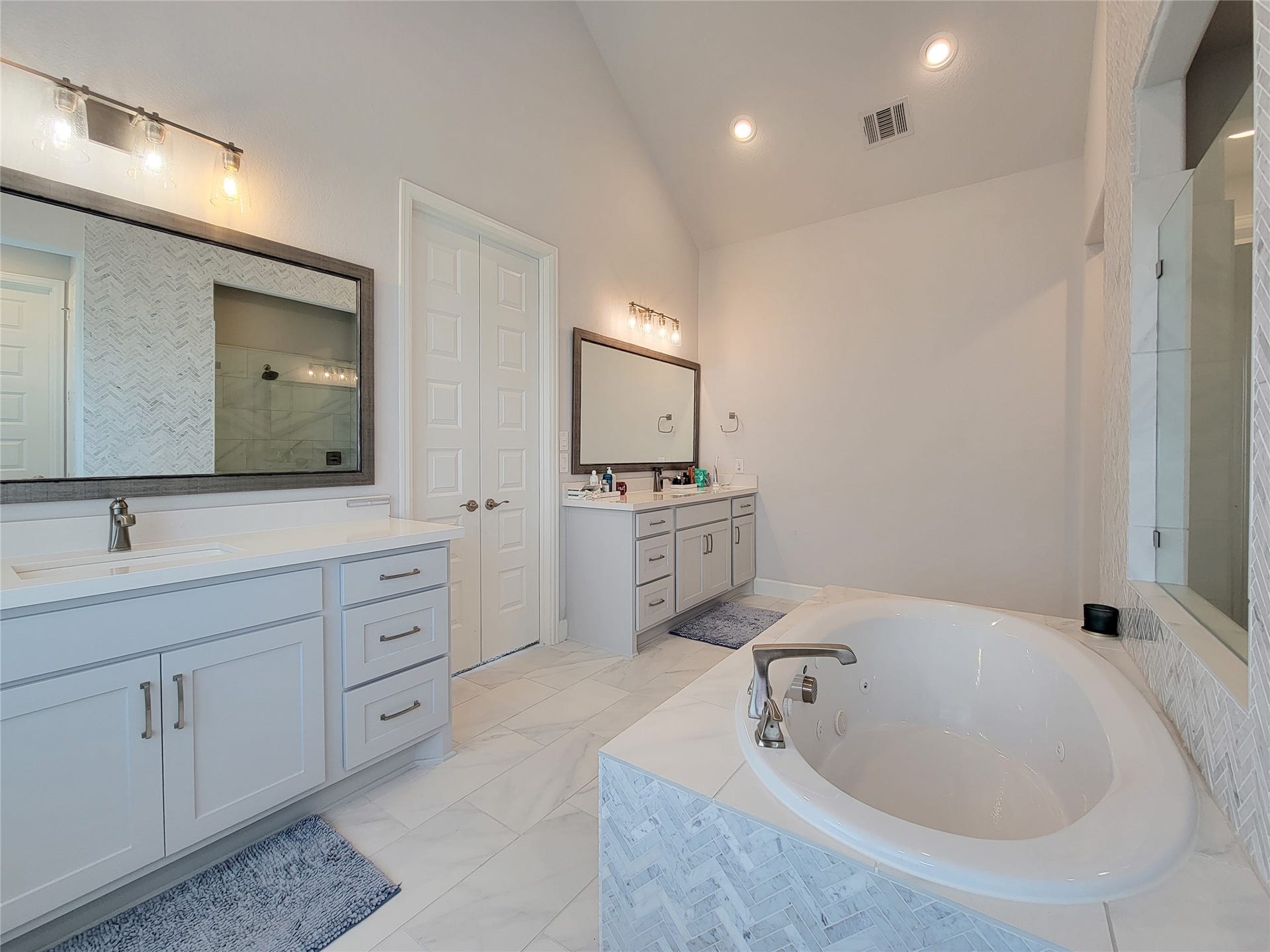 This spacious bathroom features a large soaking tub, dual vanities with modern fixtures, and ample storage. It has a bright, airy feel with elegant tile work and contemporary lighting.