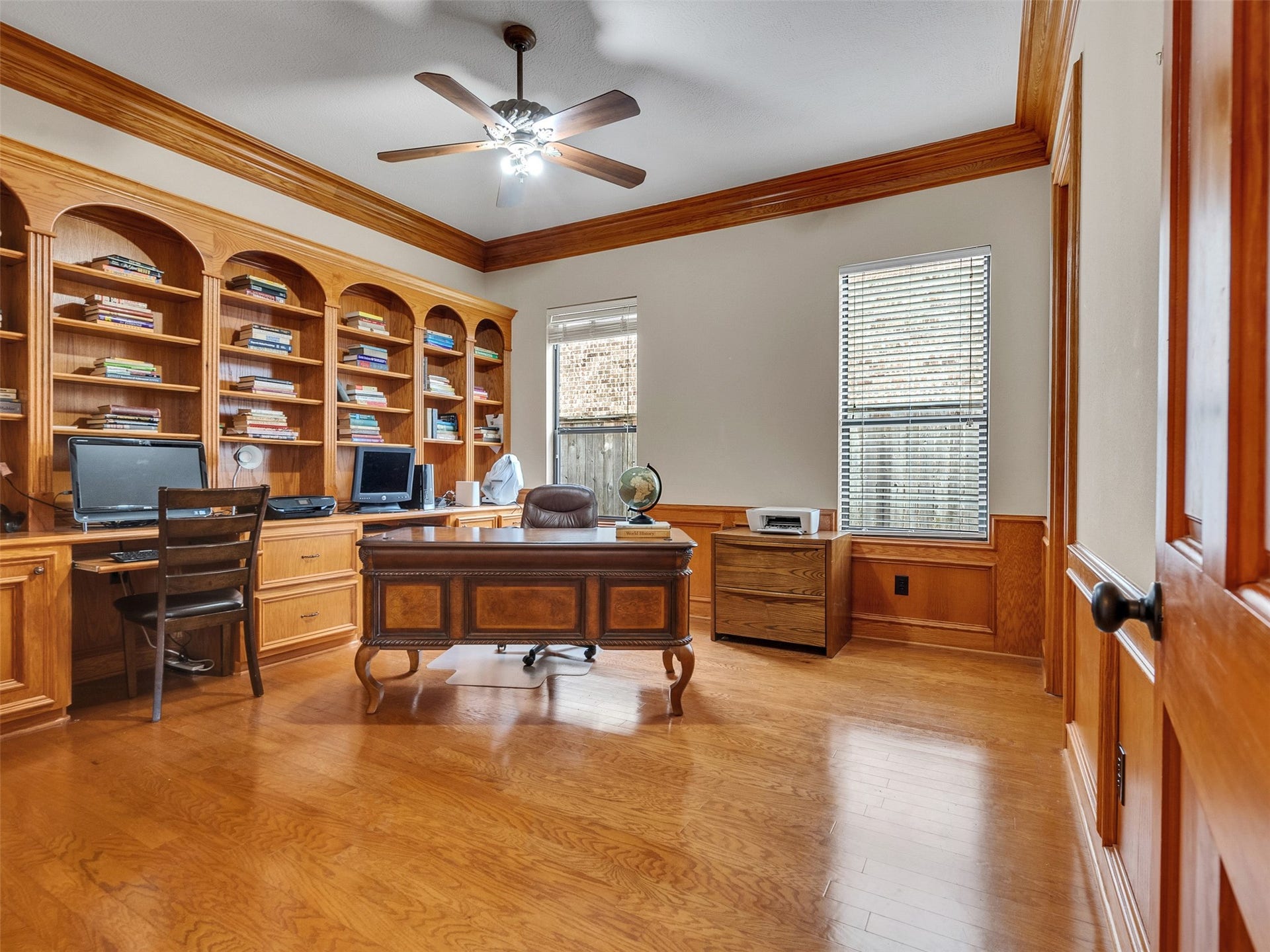 6914 Sandwedge Point Court, Spring, TX 77389 This spacious home office features elegant wood paneling, built-in bookshelves, and ample natural light from two large windows. It includes a ceiling fan and a sophisticated wood desk, creating a warm and inviting workspace.