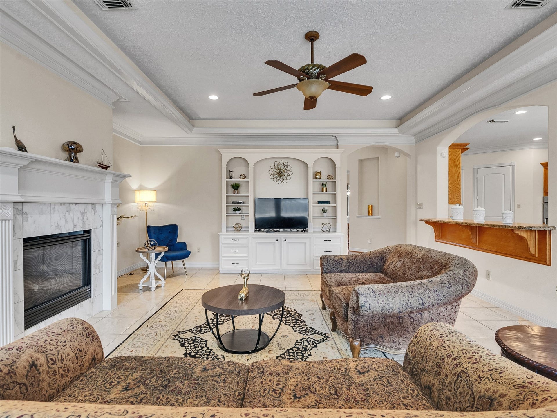6914 Sandwedge Point Court, Spring, TX 77389 This inviting living room features a cozy fireplace, built-in shelving with a TV, elegant crown molding, and a ceiling fan. The space is well-lit and includes comfortable, patterned seating and a stylish area rug, creating a warm and welcoming atmosphere.