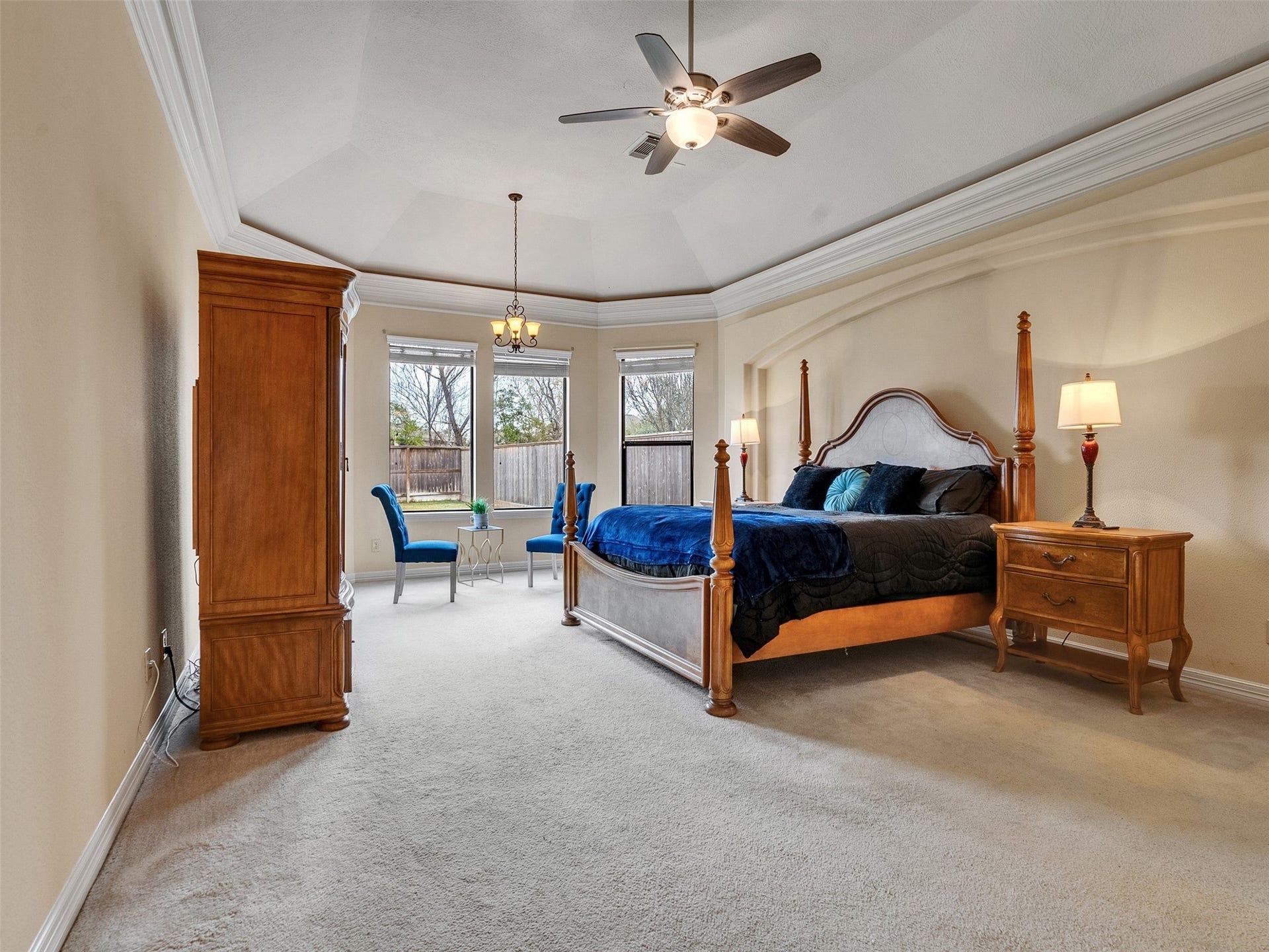 6914 Sandwedge Point Court, Spring, TX 77389 This spacious Master Bedroom features a tray ceiling with a fan, large windows providing natural light, and elegant furniture including a four-poster bed and matching wooden pieces. Cozy and inviting, it also offers a small seating area by the windows.