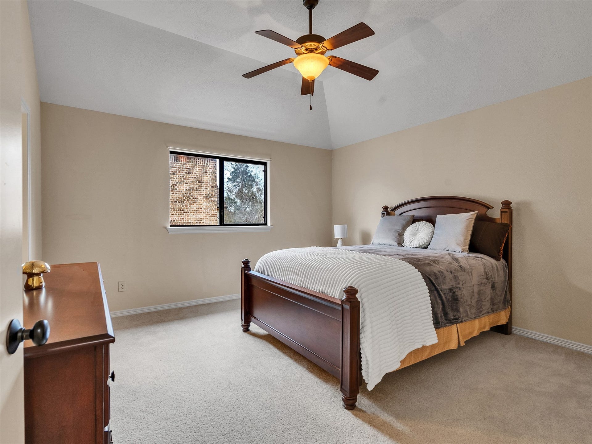 6914 Sandwedge Point Court, Spring, TX 77389 This bedroom features a vaulted ceiling with a ceiling fan, neutral walls, and carpeted flooring. It includes a window with a view and is furnished with a classic wooden bed and dresser, creating a cozy and inviting space.