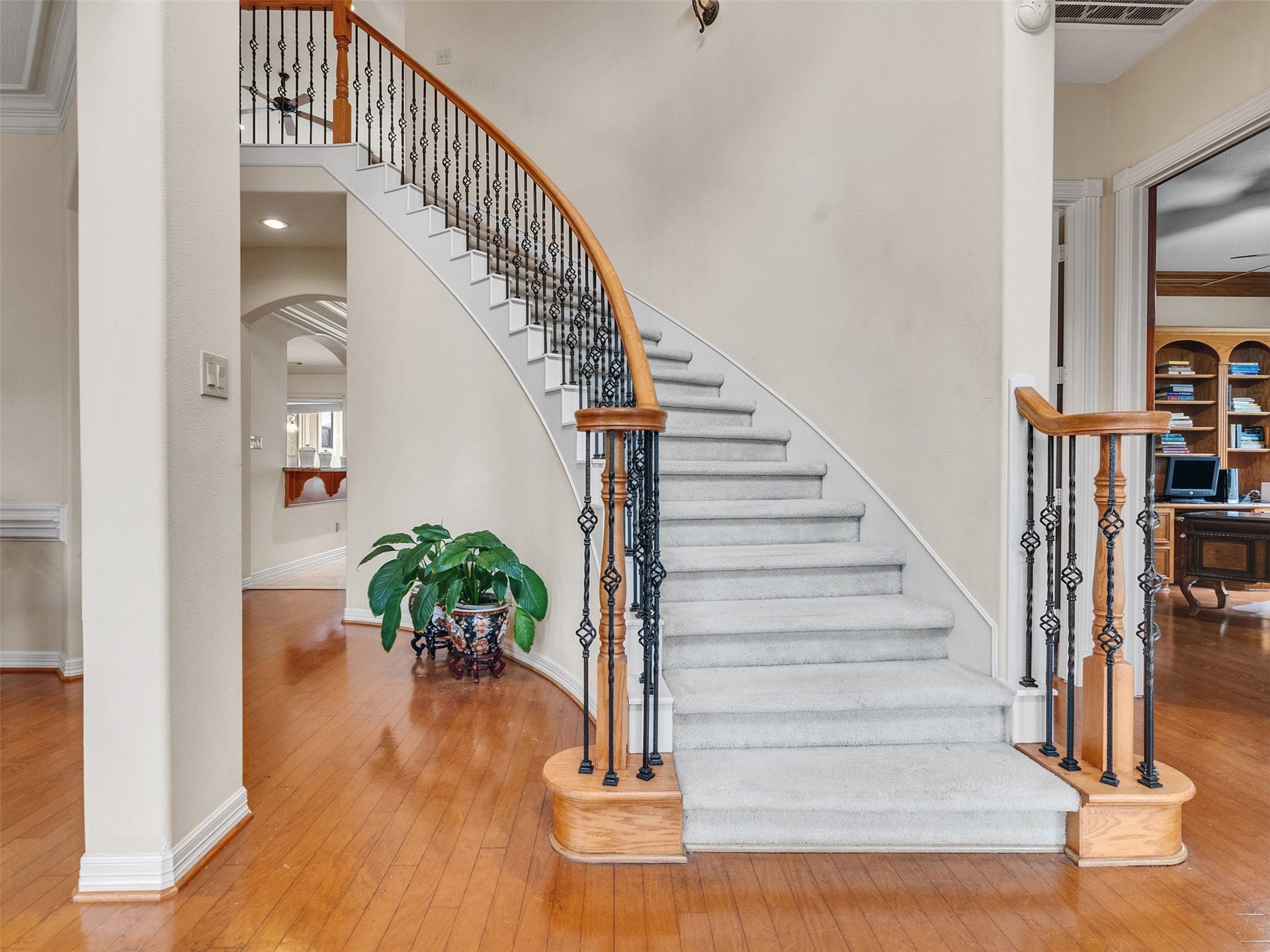 6914 Sandwedge Point Court, Spring, TX 77389 This photo showcases a welcoming entryway with a grand curved staircase featuring elegant wrought iron railings. The space is well-lit and has warm hardwood flooring, leading into a room with built-in shelves, ideal for an office or library.