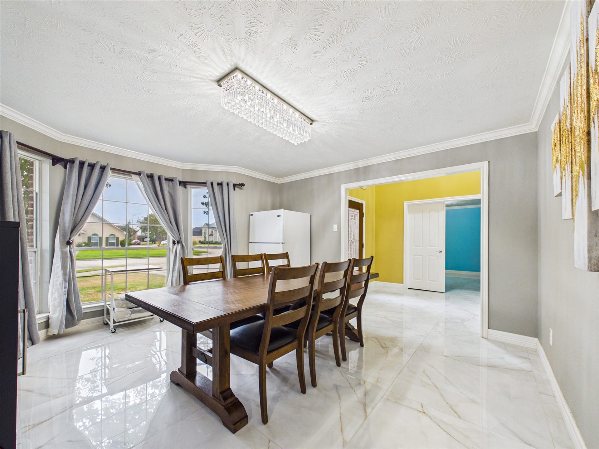 Bright dining room with a modern chandelier, large bay window, and sleek tile flooring, offering a welcoming and spacious atmosphere.