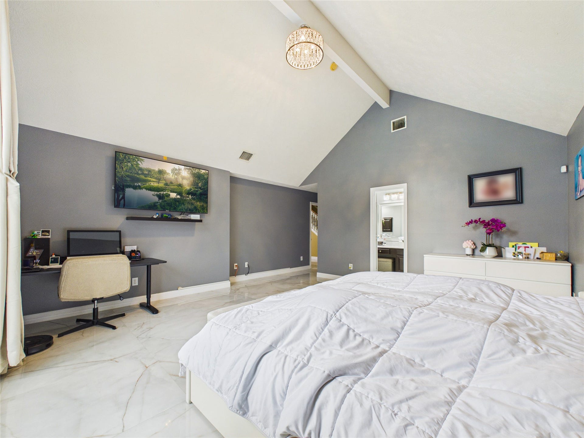Spacious bedroom with vaulted ceilings, elegant chandelier, and sleek gray walls. Features a cozy workspace, wall-mounted TV, and en-suite access. Abundant natural light enhances the modern, airy feel.