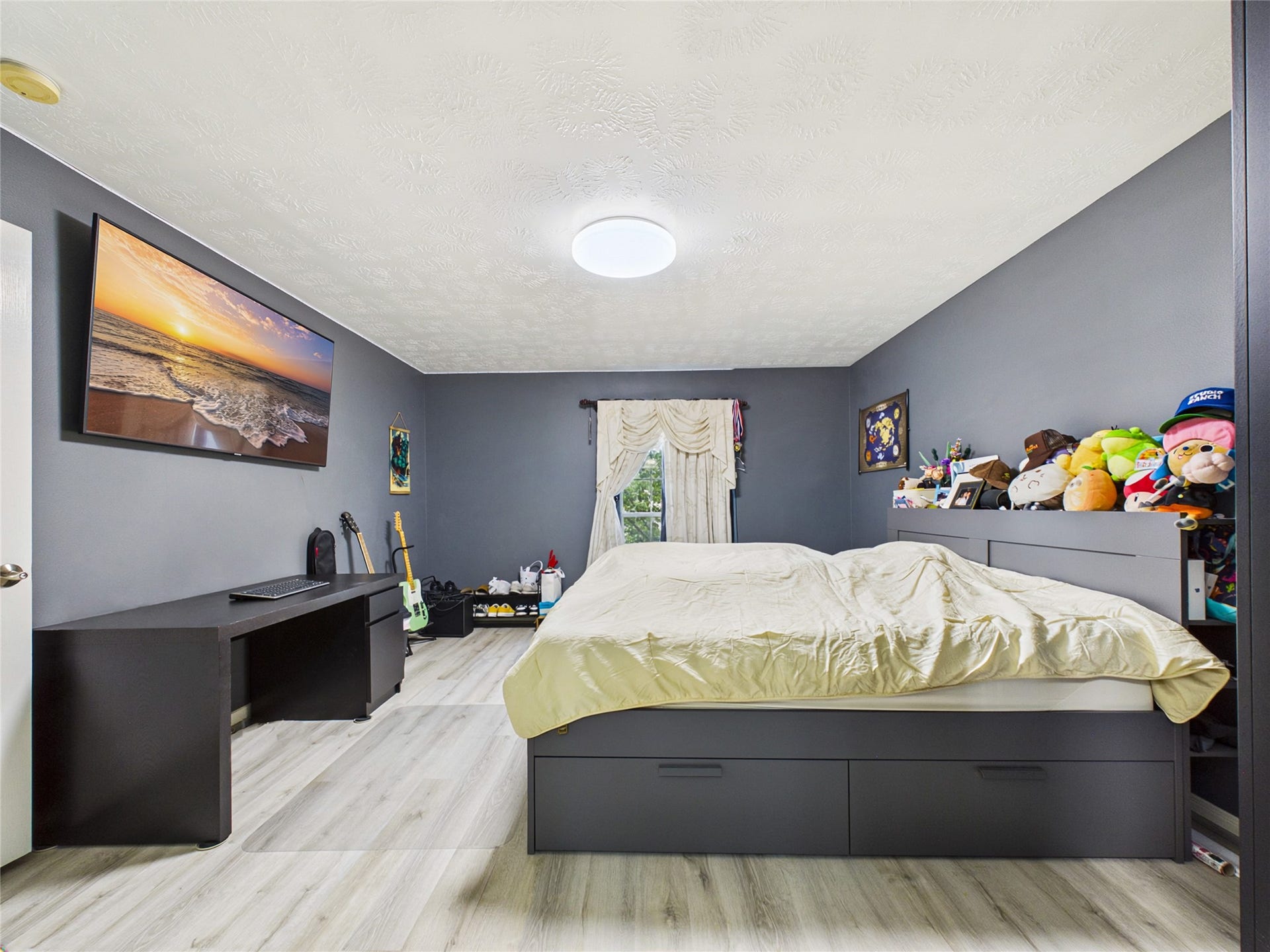 A cozy bedroom featuring sleek dark gray walls, laminate flooring, and a modern bed with storage drawers. The room is equipped with a large TV and a work desk, creating a functional and inviting space.