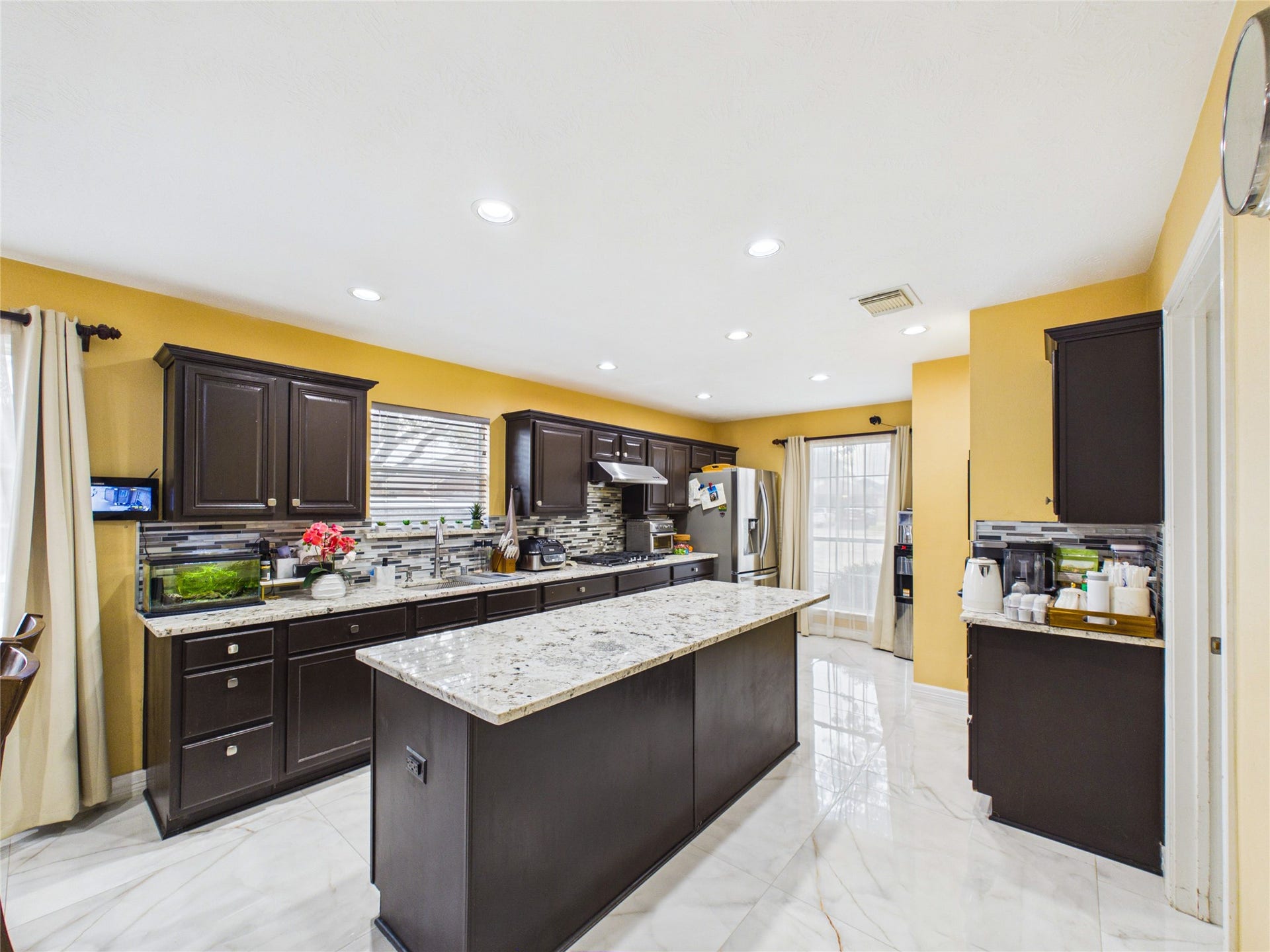 Spacious kitchen with elegant dark wood cabinets, granite countertops, and modern stainless steel appliances, accented by vibrant yellow walls and natural light from large windows.