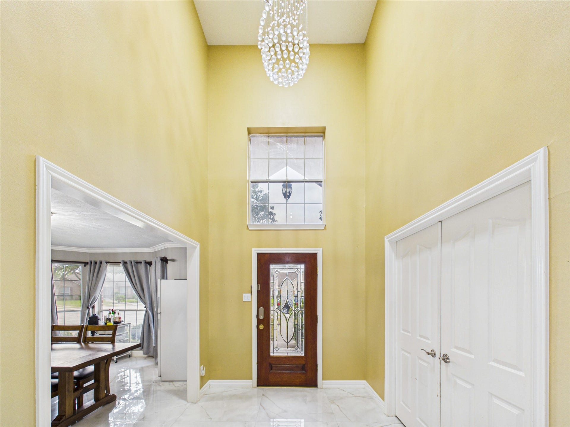 Elegant foyer with high ceilings, a stunning chandelier, and a decorative glass front door, leading into a bright dining area with large windows.
