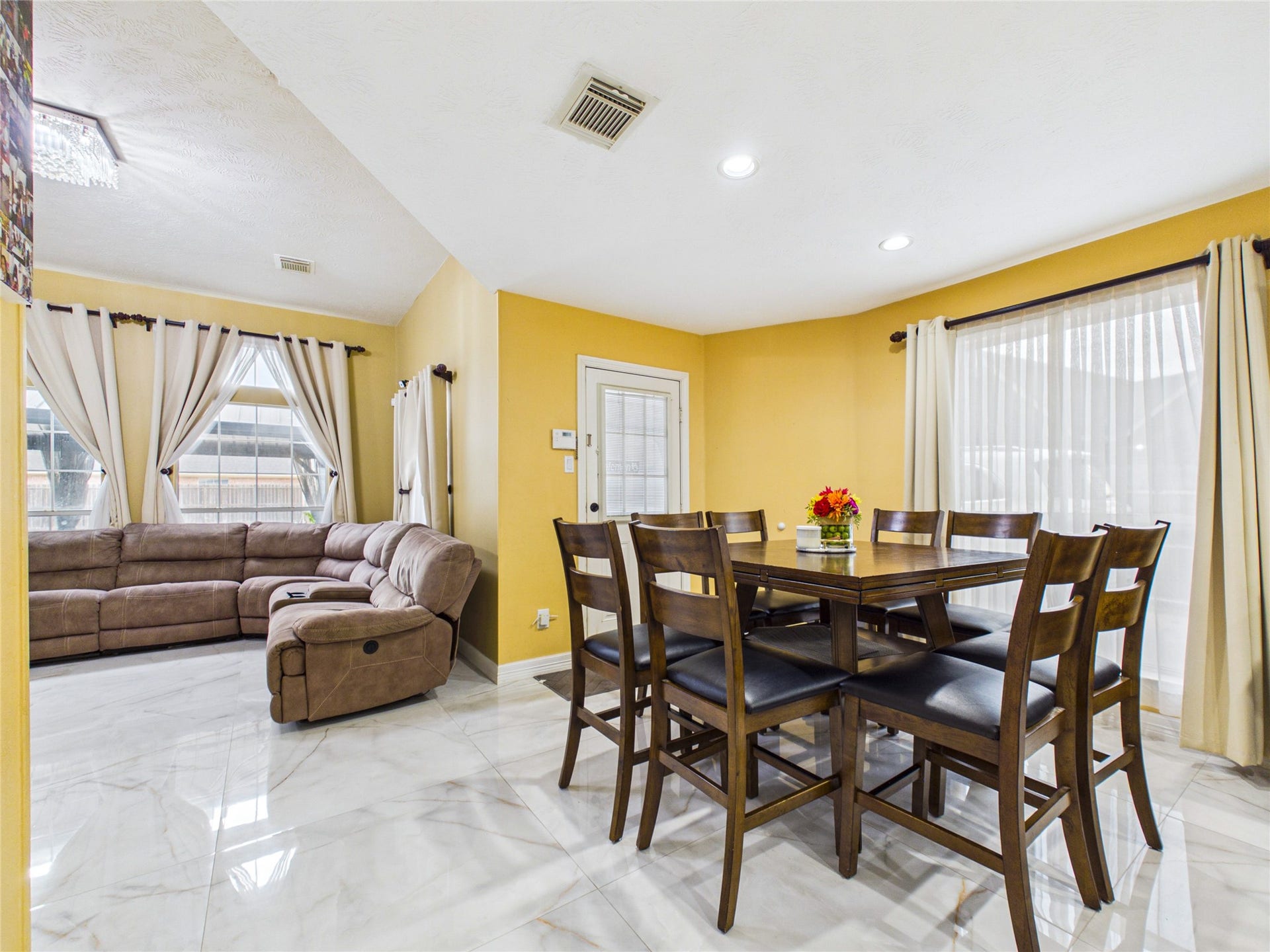 Bright and inviting open-concept living and dining area features elegant marble floors, a cozy sectional sofa, and ample natural light from large windows with sheer curtains.