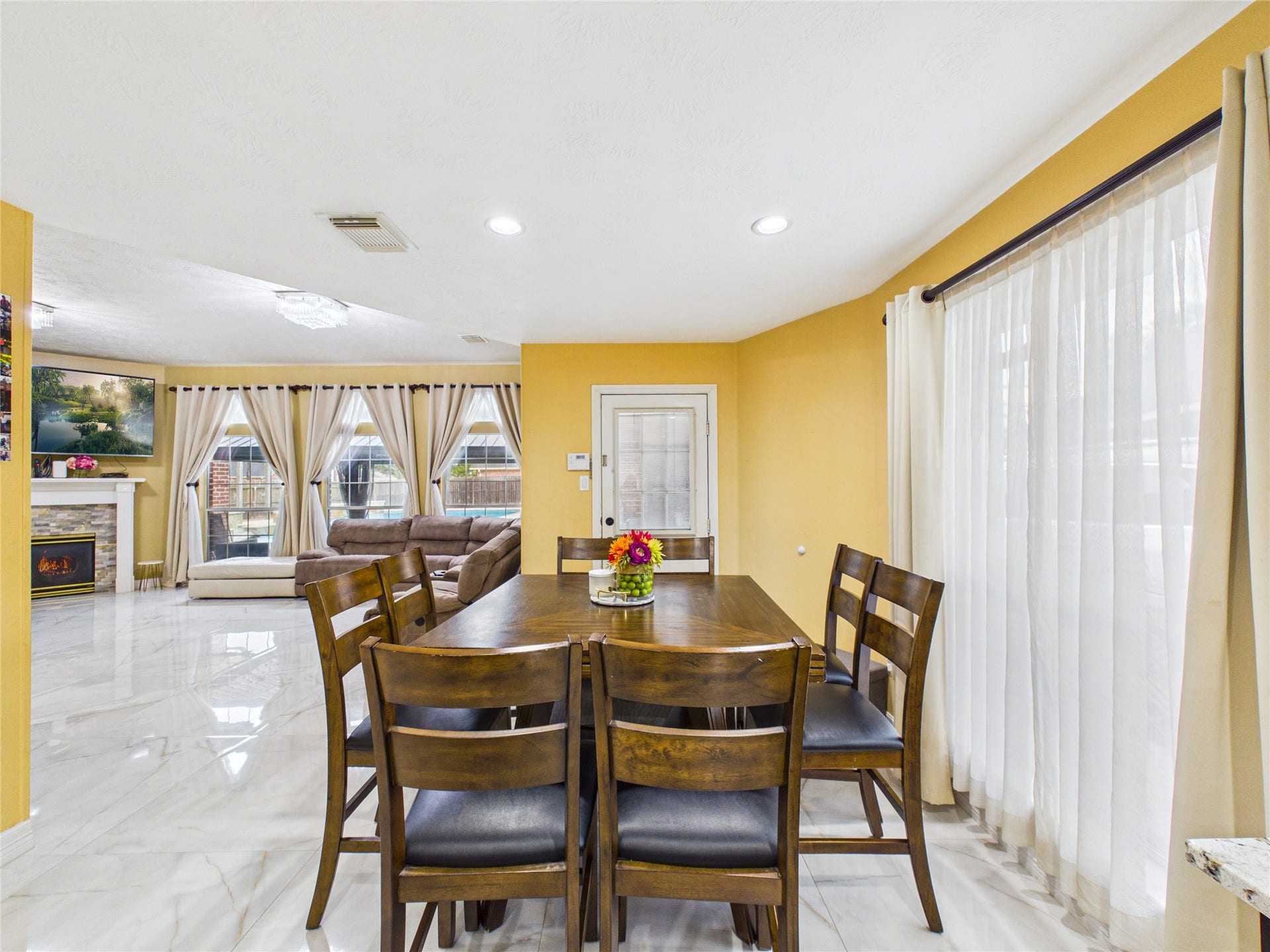 Bright and inviting dining area with elegant tile flooring, warm yellow walls, and large windows offering abundant natural light, seamlessly flowing into a cozy living space with a fireplace.