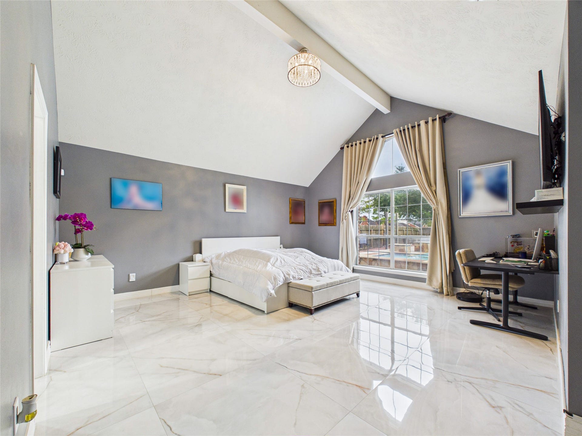 Spacious bedroom with vaulted ceilings, modern decor, and abundant natural light from large windows, featuring sleek tile flooring and a cozy workspace.