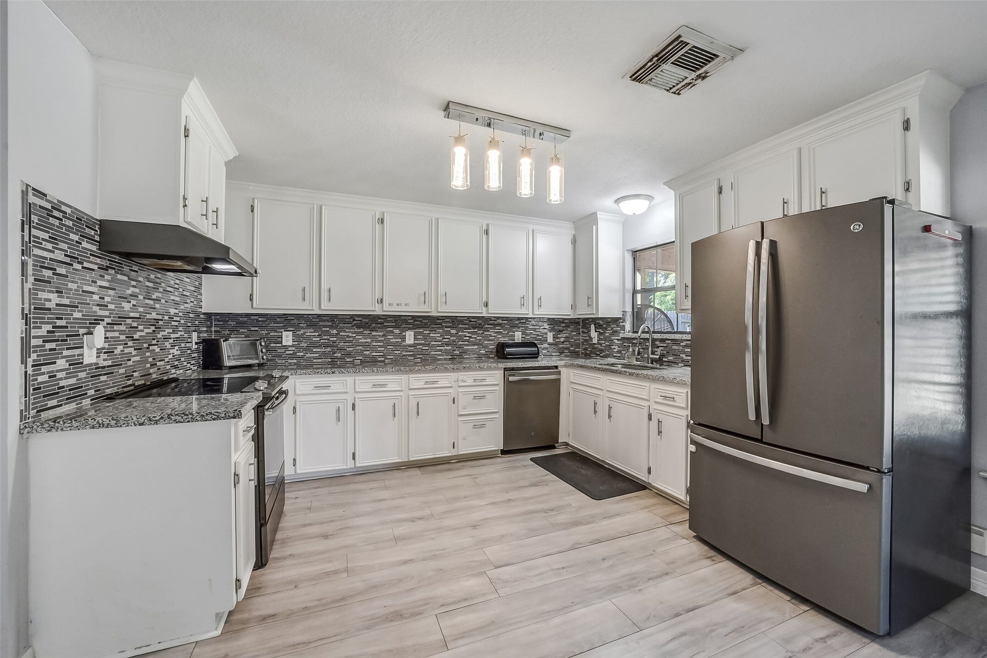 22911 Keith Drive, New Caney, TX 77357 This bright kitchen features abundant white cabinetry, granite countertops, and a striking mosaic tile backsplash. Stainless steel appliances, contemporary lighting, and wood-look flooring complement the space, while a window above the sink welcomes natural light.