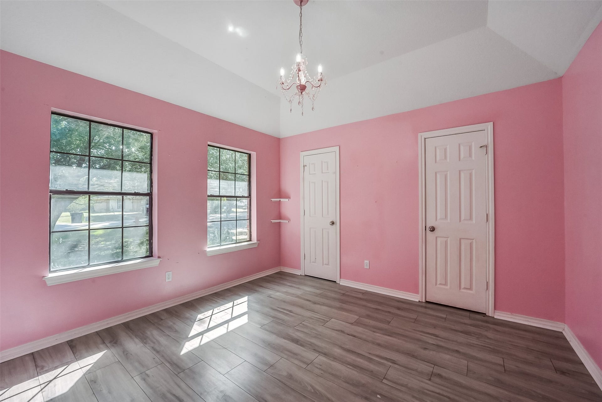 22911 Keith Drive, New Caney, TX 77357 This charming secondary bedroom features high ceilings, a decorative chandelier, and two large windows that fill the space with natural light. Pink walls, crisp white trim, and wood-look flooring create a bright, inviting atmosphere, while corner shelves offer a thoughtful decorative touch.
