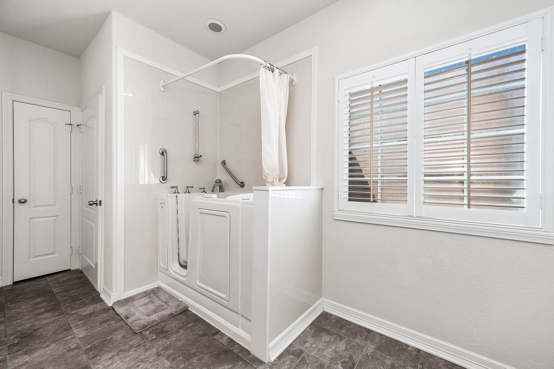Spacious bathroom featuring a convenient walk-in tub, elegant tile flooring, and bright natural light through the shuttered window.