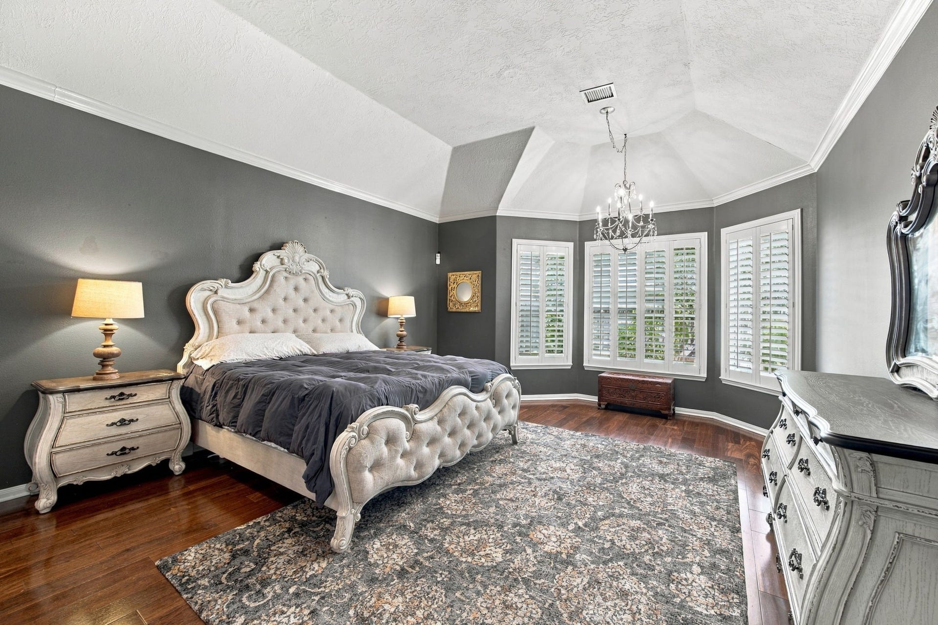 2417 La Rochelle Court, Seabrook, TX 77586 Elegant primary bedroom featuring rich hardwood floors, and a stylish chandelier. Natural light pours in through large windows with plantation shutters, creating a serene and inviting atmosphere.