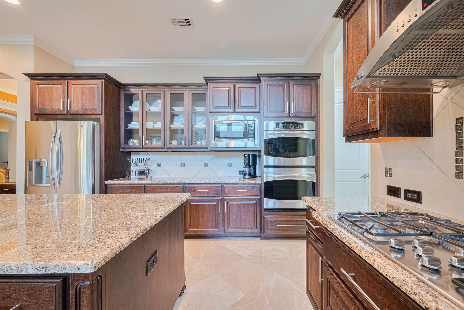 4722 Burclare Court, Sugar Land, TX 77479 Detailed culinary view showcasing the five-burner gas cooktop, vent hood, stacked ovens, glass-front cabinets, and stainless steel refrigerator.