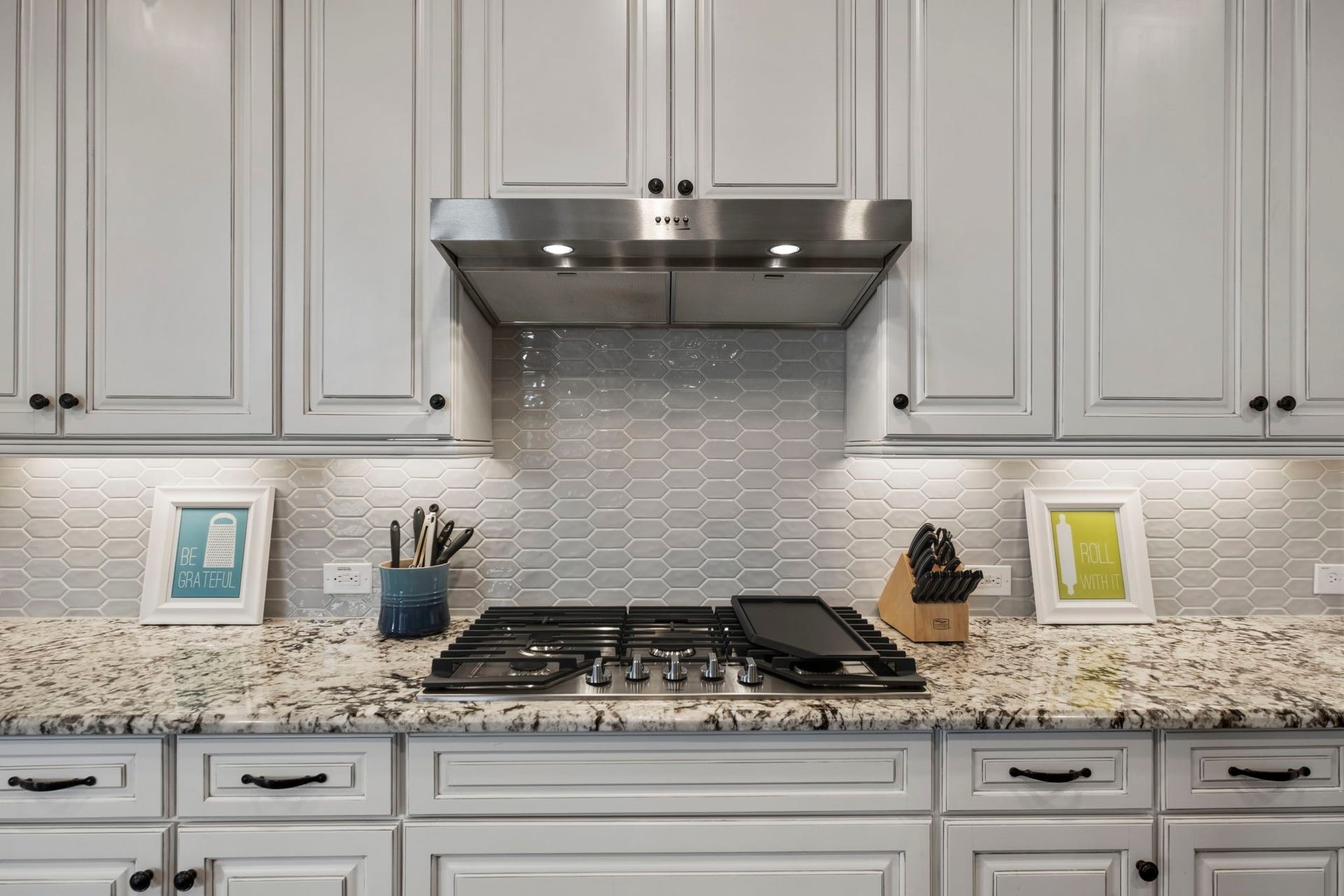 19314 Trotting Green Trail, Tomball, TX 77377 This kitchen features elegant white cabinetry with dark knobs, a modern stainless steel range hood, and a sleek gas stove set against a stylish hexagonal tile backsplash. The granite countertops add a touch of luxury, complemented by under-cabinet lighting.