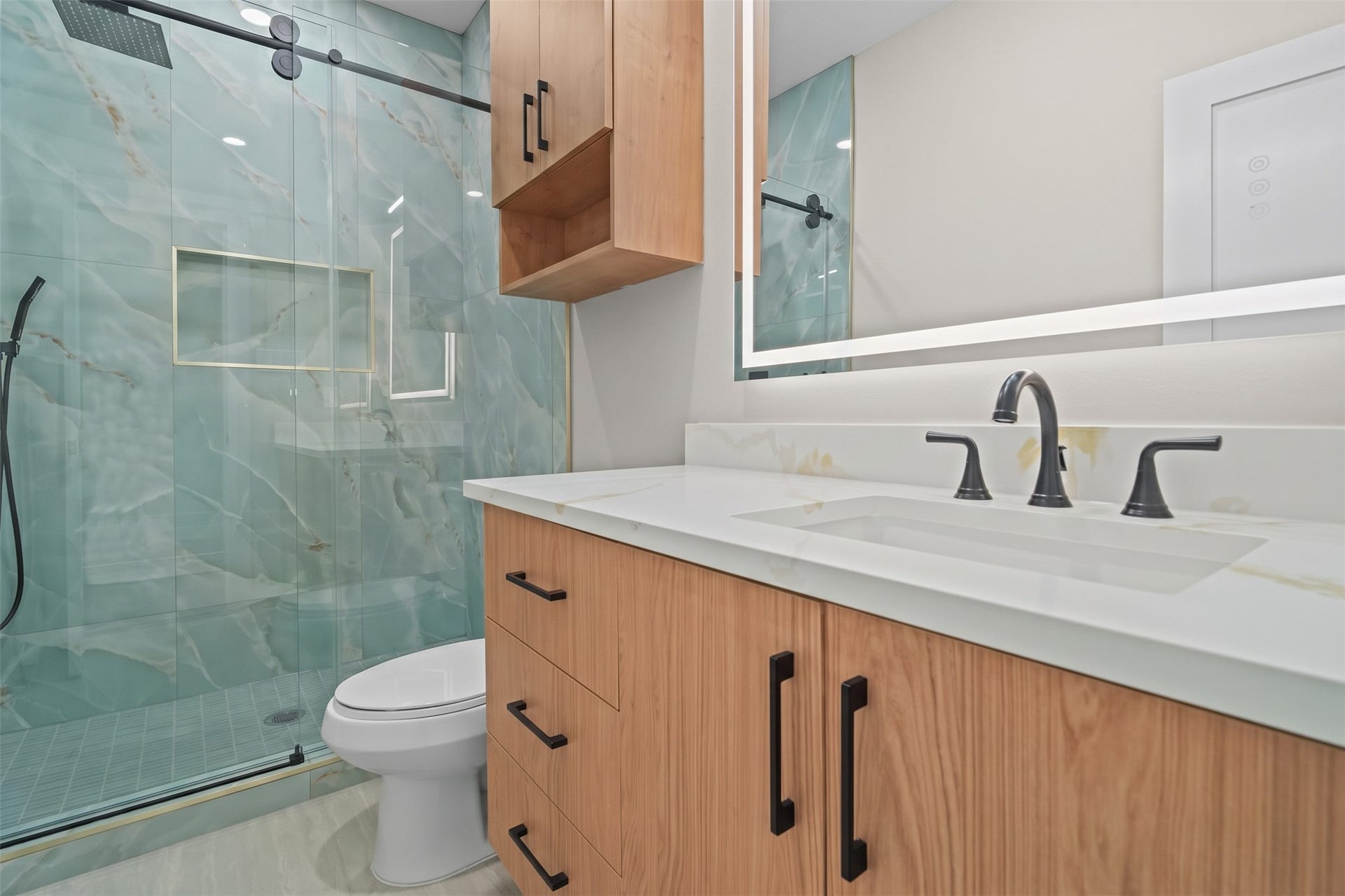 This bathroom offers a spacious single-sink vanity and custom overhead storage. A frameless glass walk-in shower with full wall tiling and a handheld sprayer creates a spa-like feel in a compact footprint.