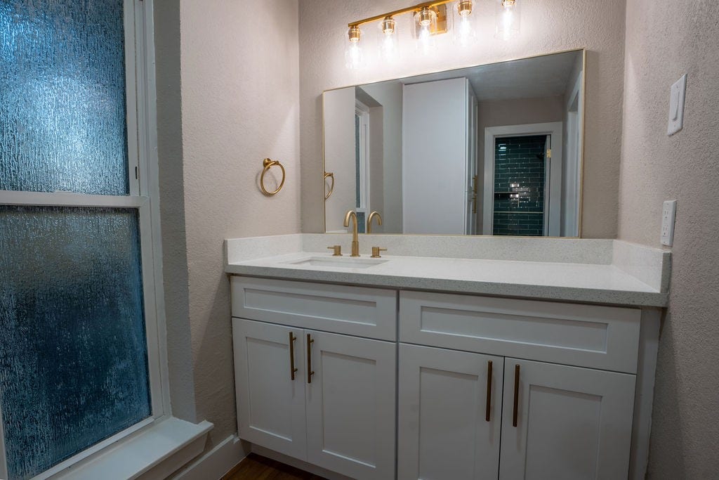 9856 Oriole, Conroe, TX 77385 Elegant bathroom featuring a modern vanity with sleek white cabinetry, gold fixtures, and a large mirror under a stylish light fixture.