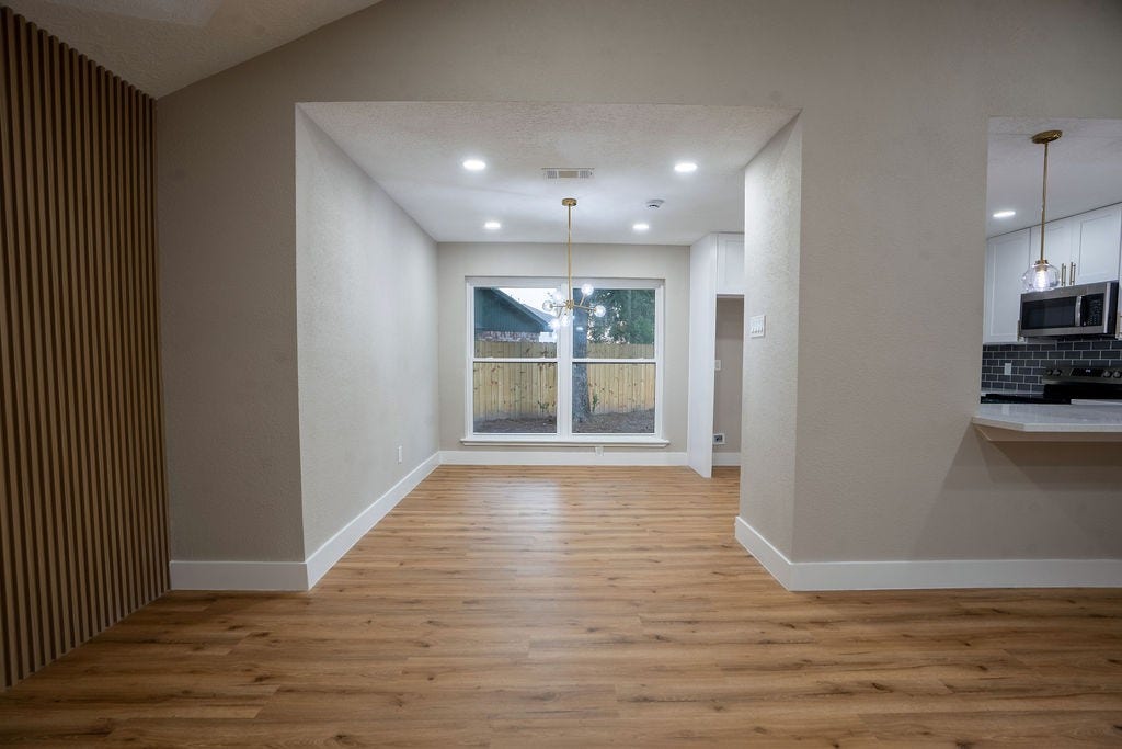 9856 Oriole, Conroe, TX 77385 Elegant open-concept living space featuring warm vinyl flooring, modern lighting, and a large window allowing natural light to fill the room. Adjacent kitchen area boasts sleek cabinetry and contemporary finishes.
