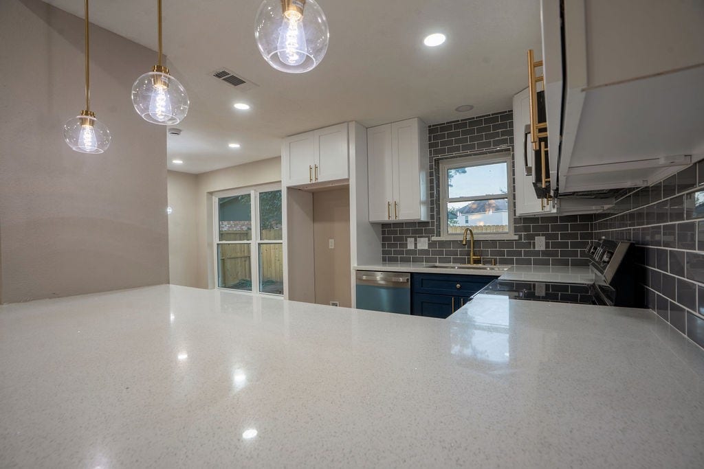 9856 Oriole, Conroe, TX 77385 Sleek modern kitchen featuring glossy quartz countertops, elegant pendant lighting, and stylish gray subway tile backsplash, complemented by stainless steel appliances and a charming window view.