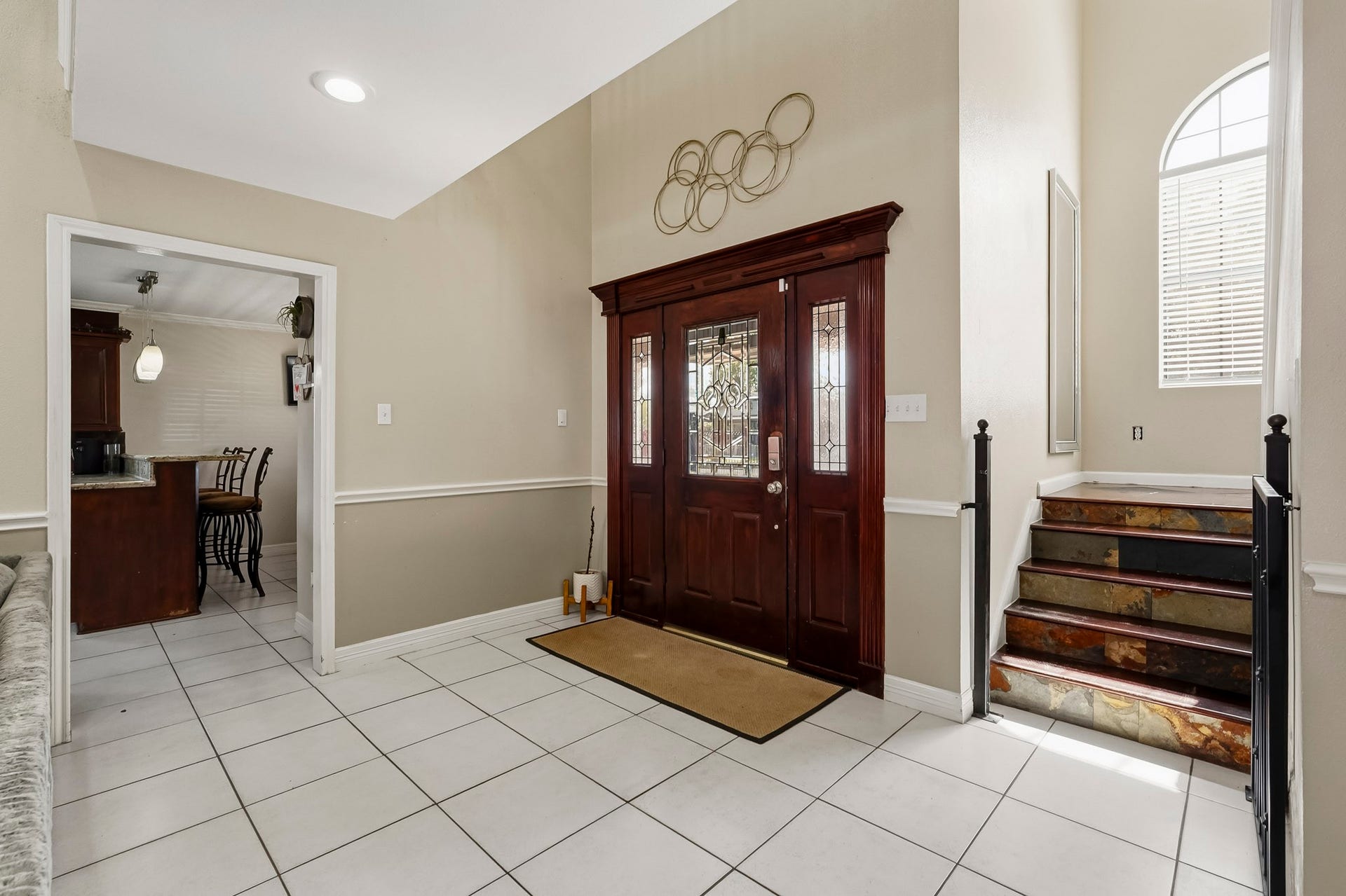 1207 9th Street, Galena Park, TX 77547 Elegant entryway with a rich wooden front door featuring decorative glass, leading to a bright, tiled foyer. An inviting staircase with wrought iron railings adds charm, while an adjacent doorway opens to a cozy kitchen area.