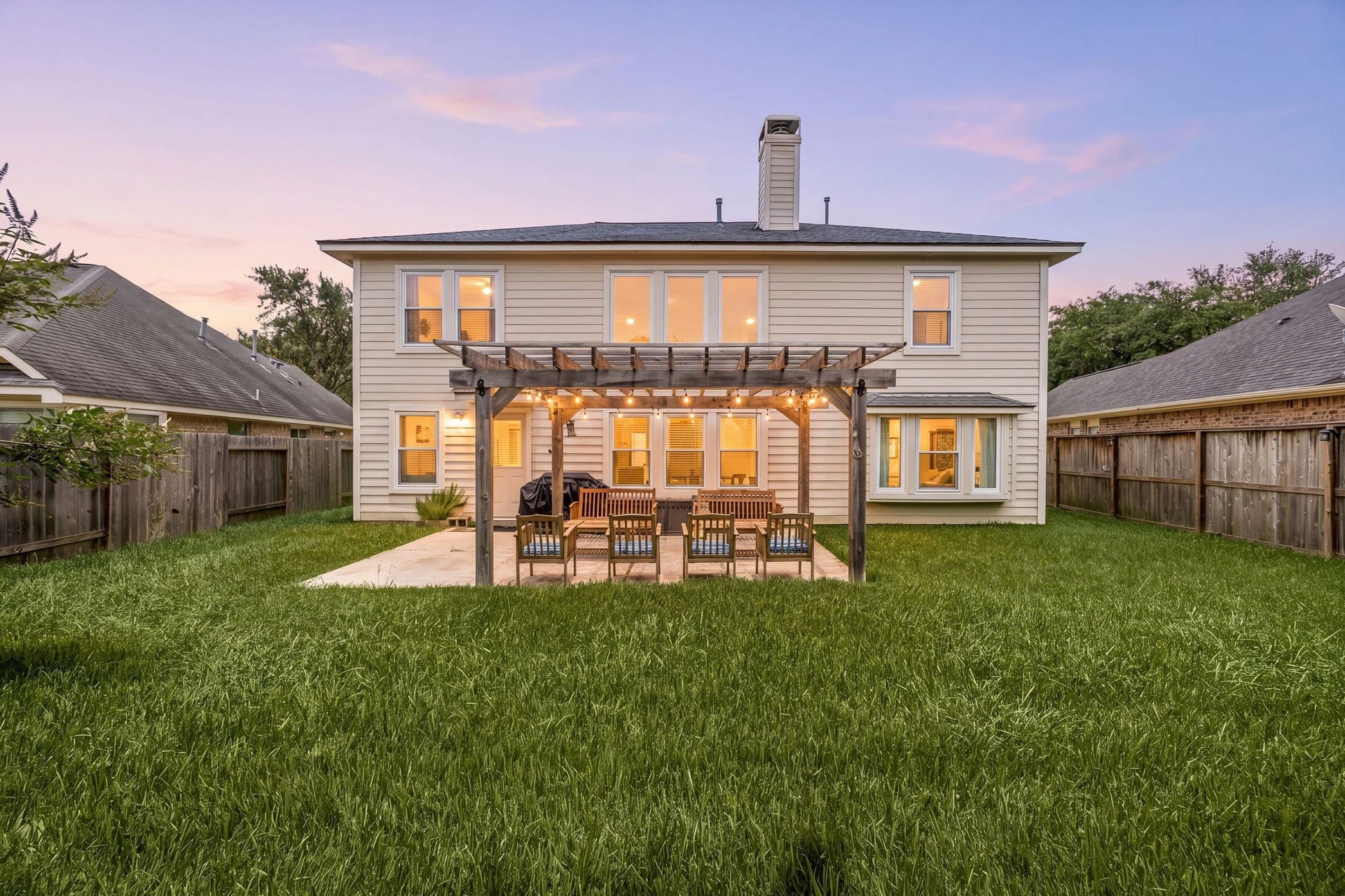 3915 Eastland Lake Drive, Richmond, TX 77406 Evening lighting highlights the expansive fully fenced backyard layout, showing the rear exterior of the home, a manicured grass lawn, and the custom wood pergola detailed with outdoor string lighting.