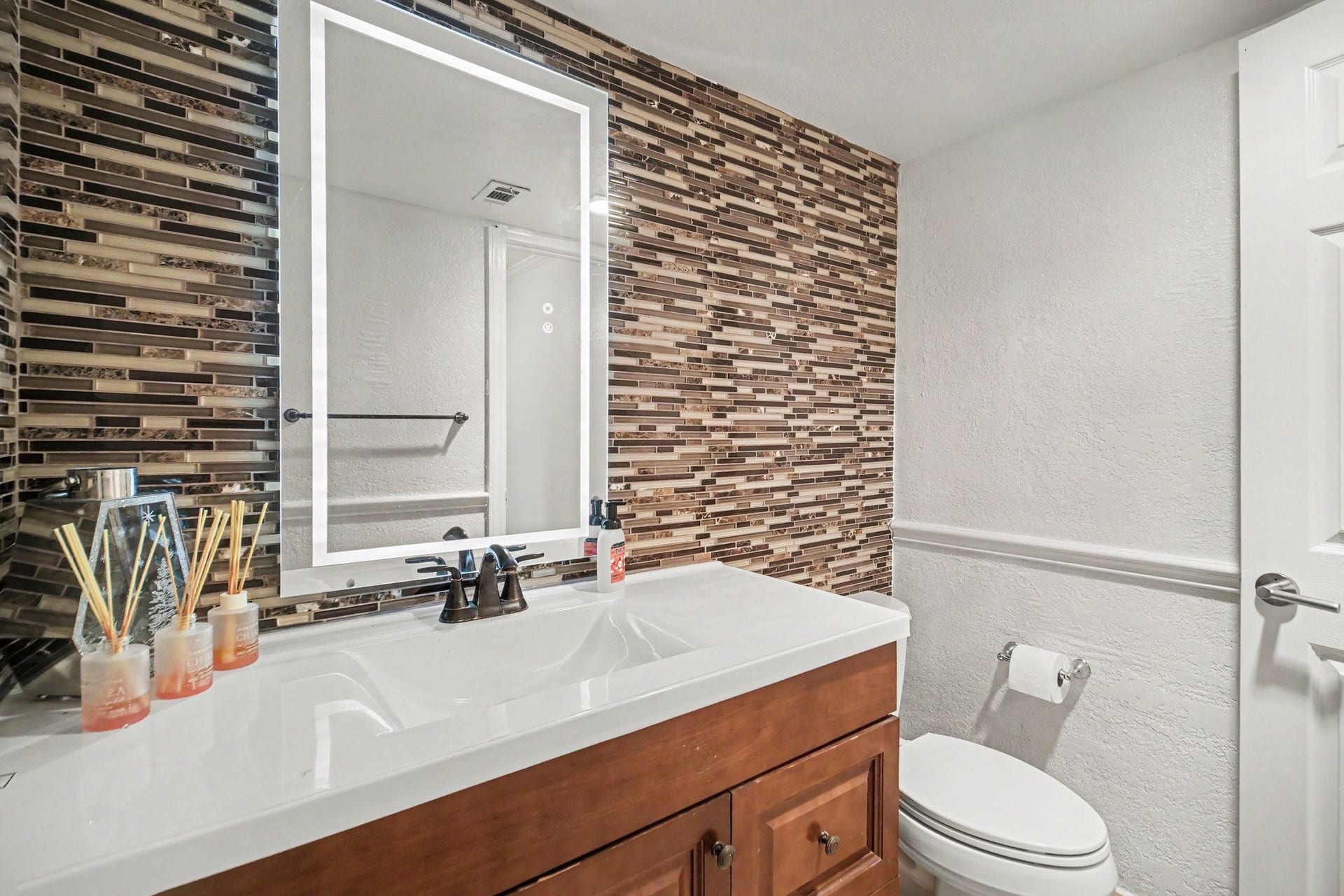 Sleek and stylish half bath featuring a modern mosaic accent wall, illuminated vanity mirror, and elegant fixtures, perfect for guests.