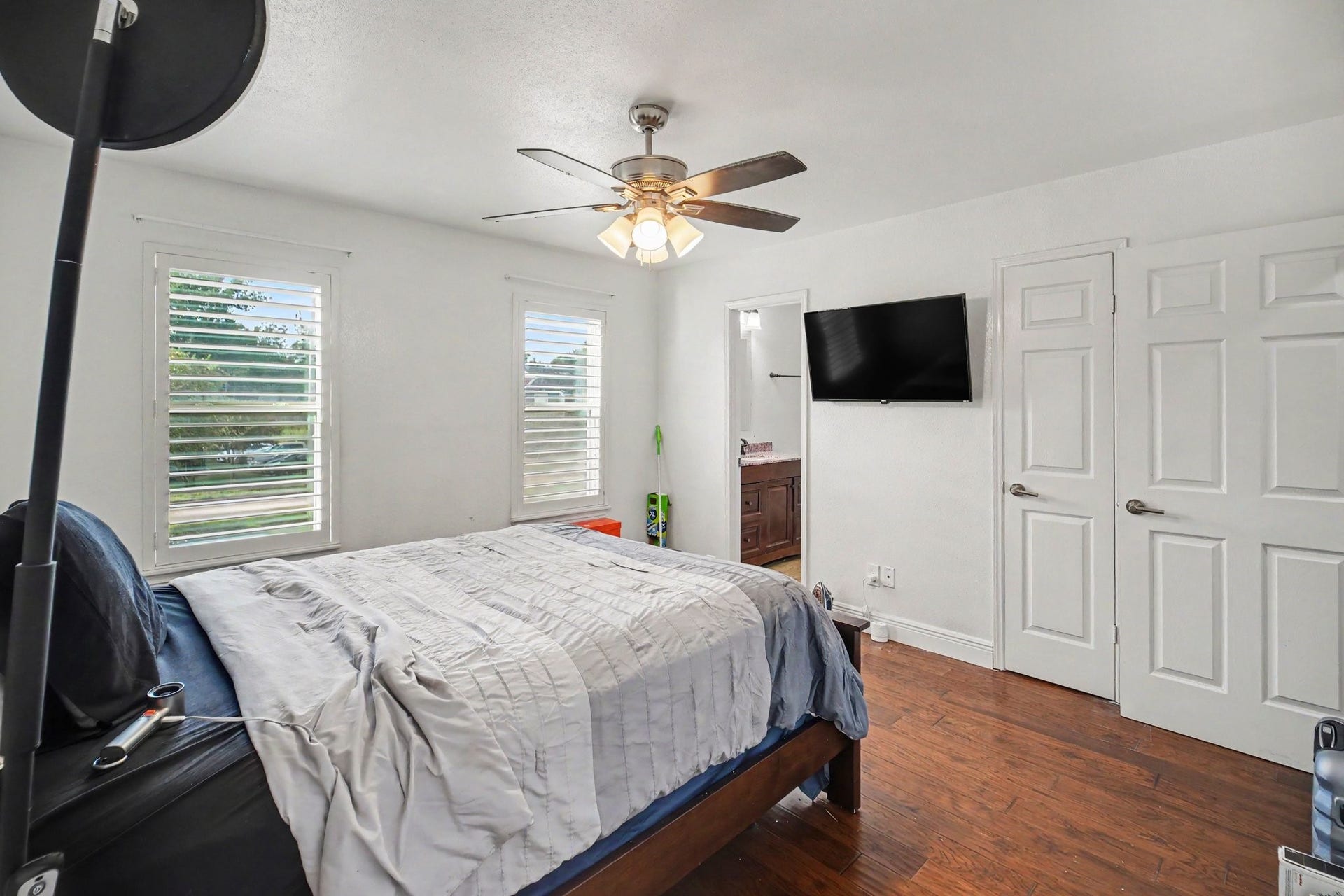 Bright and welcoming, the second bedroom features rich hardwood floors, abundant natural light from dual windows, and modern amenities like a ceiling fan and wall-mounted TV.