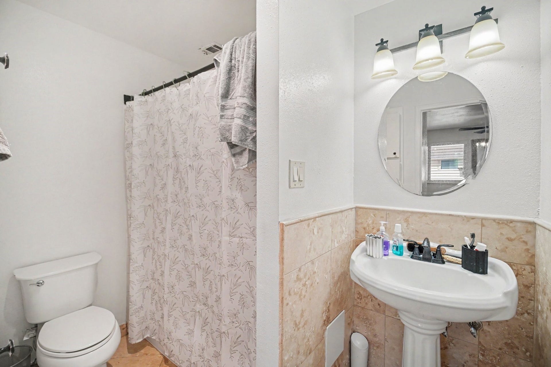 Charming bathroom with a pedestal sink, elegant round mirror, and modern lighting. The shower area features a stylish curtain, and there are tasteful tile accents throughout.