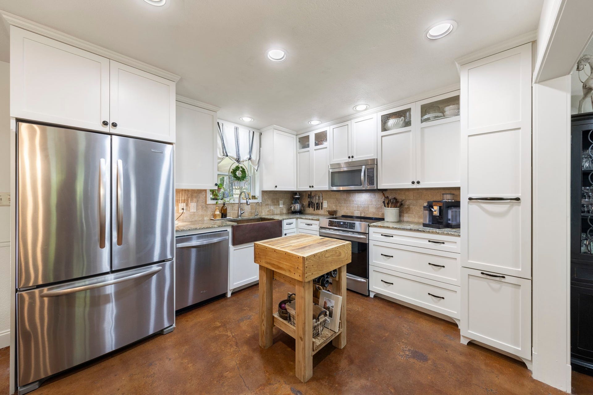 The kitchen showcases crisp white cabinetry, stainless steel appliances, a farmhouse sink, abundant storage, and a movable butcher block island that provides additional prep space while maintaining an open, functional layout.