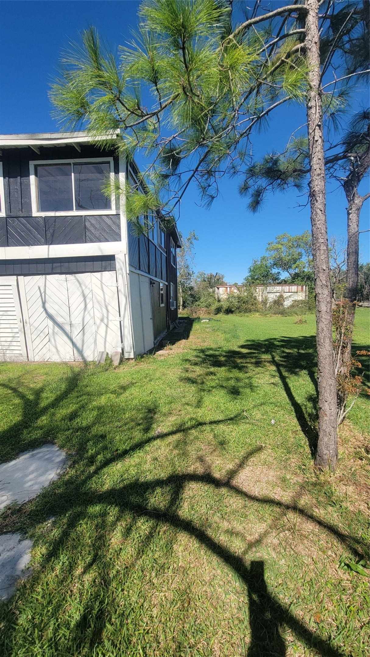 12018 Sandpiper Road, Angleton, TX 77515 This photo showcases a grassy side yard adjacent to a two-story home with a screened upper section, surrounded by trees, under a clear blue sky.