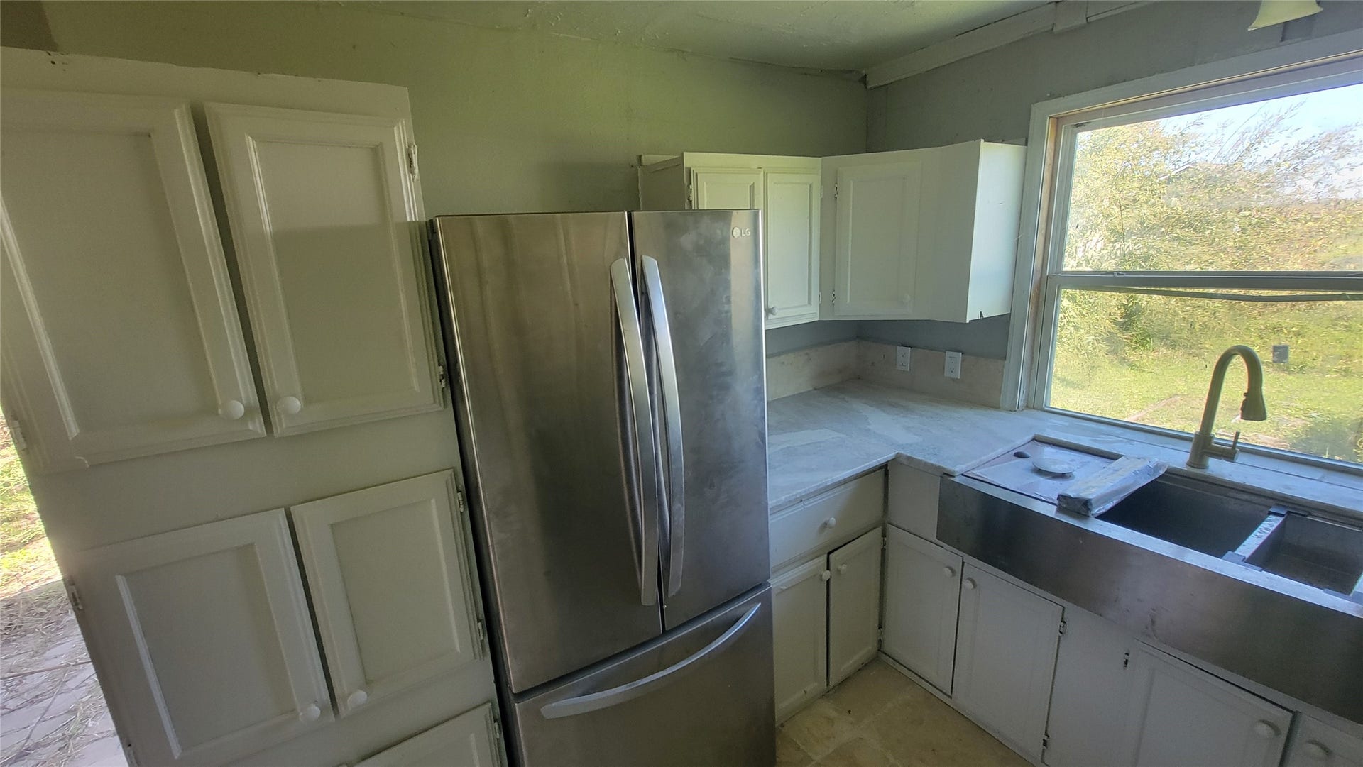 12018 Sandpiper Road, Angleton, TX 77515 This kitchen features white cabinetry, a stainless steel refrigerator, and a large window providing natural light. The space includes a double sink with a view of the greenery outside.