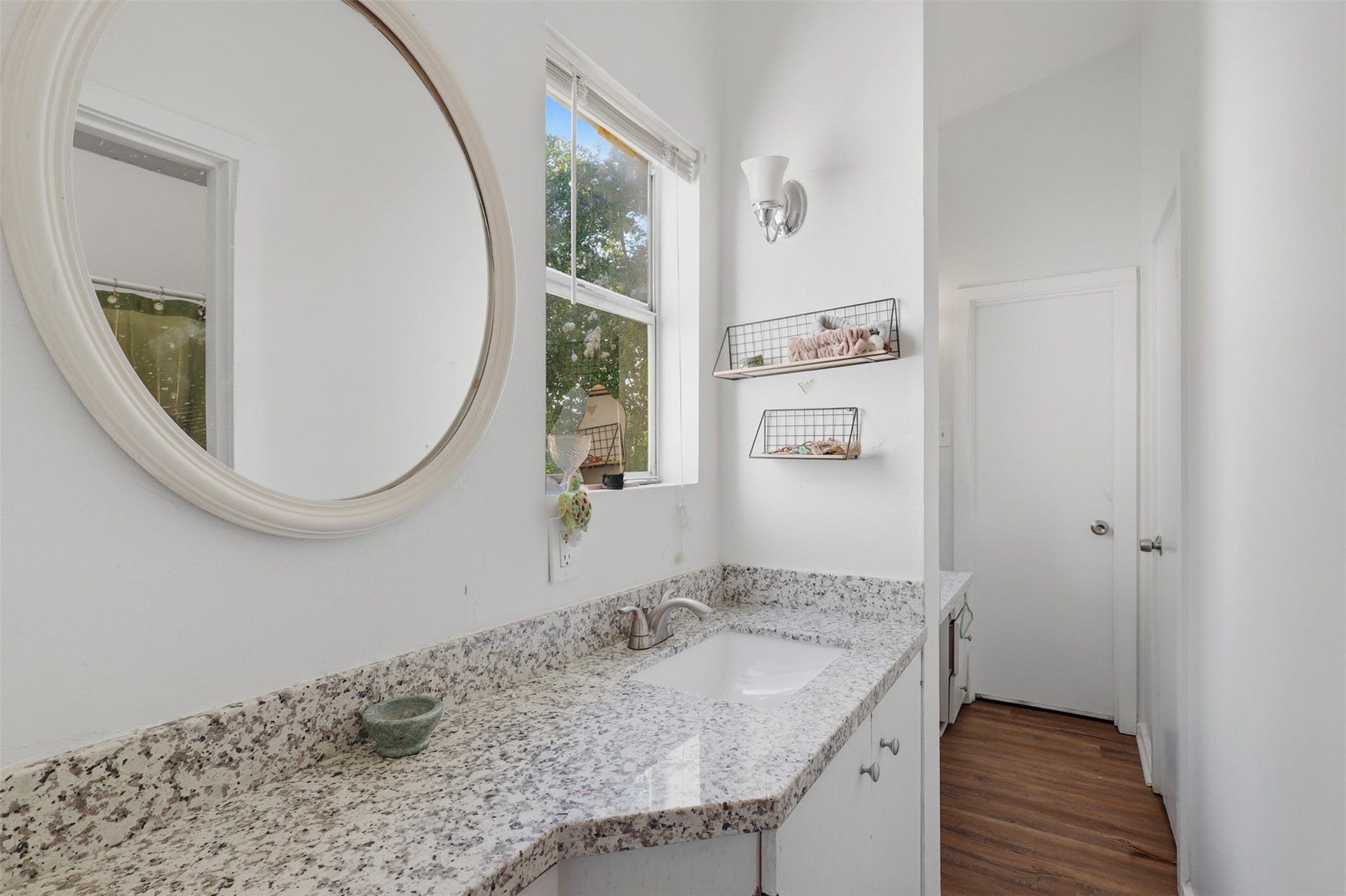 Premium granite countertop pair with an elegant round statement mirror and a bright side window in this private ensuite setup.