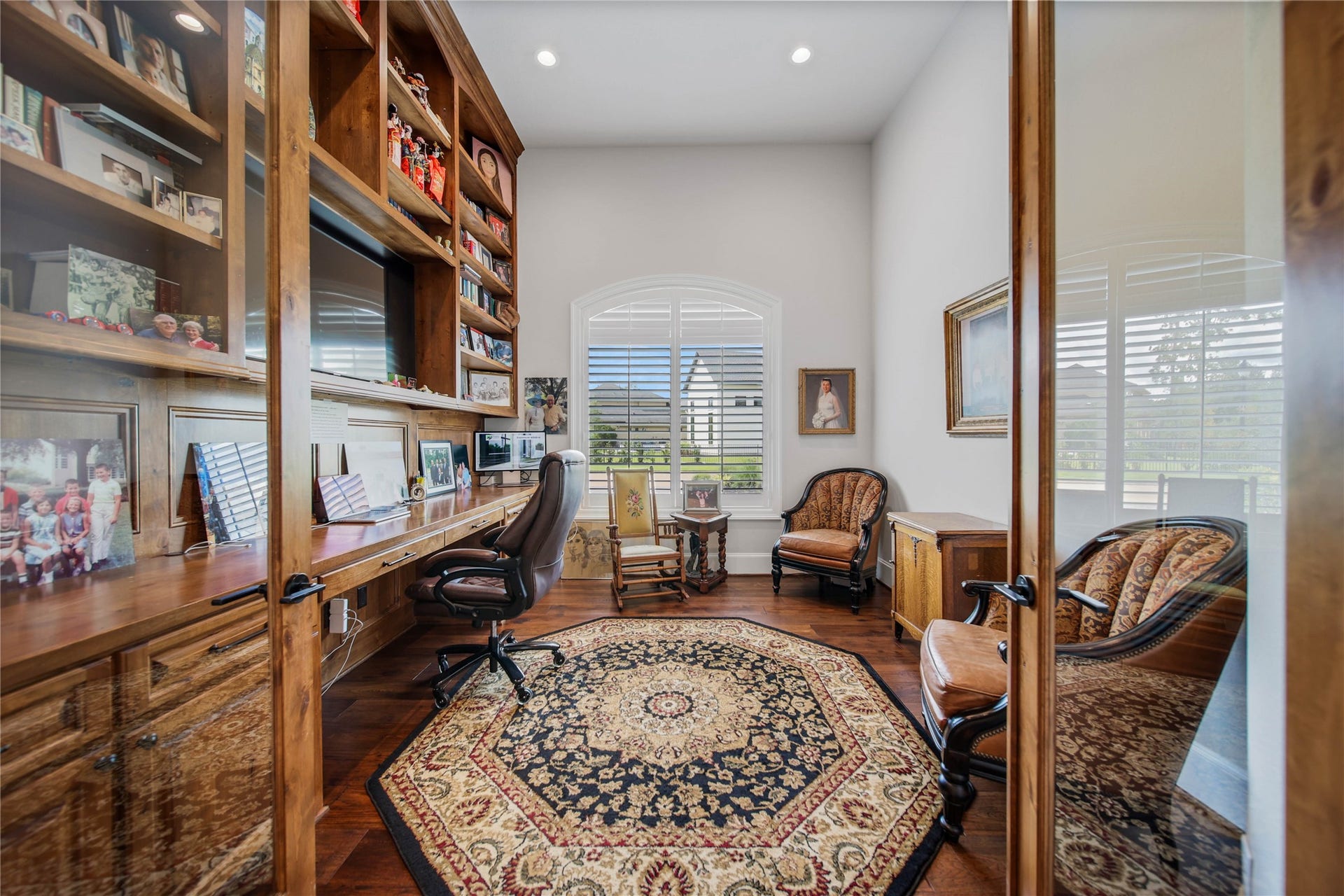 24718 Shadow Pond Drive, Spring, TX 77389 Elegant home office featuring rich wooden built-in shelving, large desk space, and plush seating, complemented by a decorative rug and abundant natural light through a large window with plantation shutters.